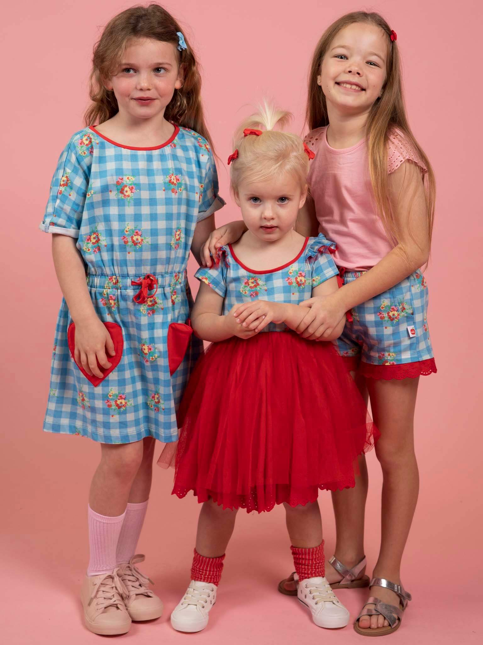 Three young girls wearing matching blue gingham outfits with a pink background