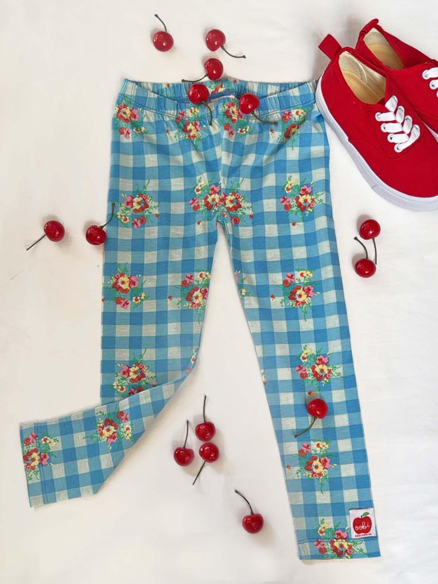 Floral Gingham Jersey Leggings by Floral Gingham with a blue and white vintage floral gingham print are laid flat, scattered cherries as decoration, and red lace-up sneakers in the top right corner.