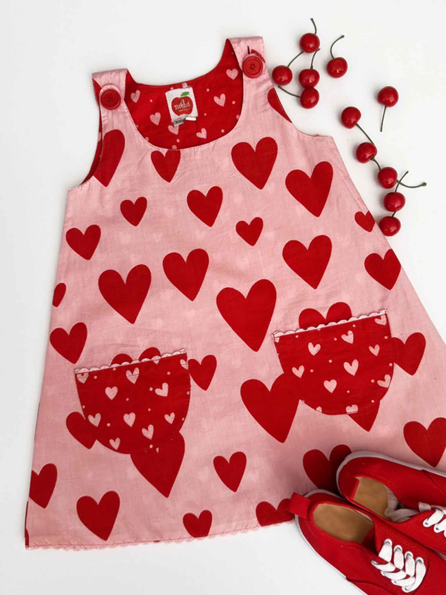 The Big Hearts "Big Heart Gracie Reversible Dress" is a pink cotton girls' smock dress with red heart patterns, two front pockets, red strap buttons, and is styled with red shoes and cherry-like decorations beside it.
