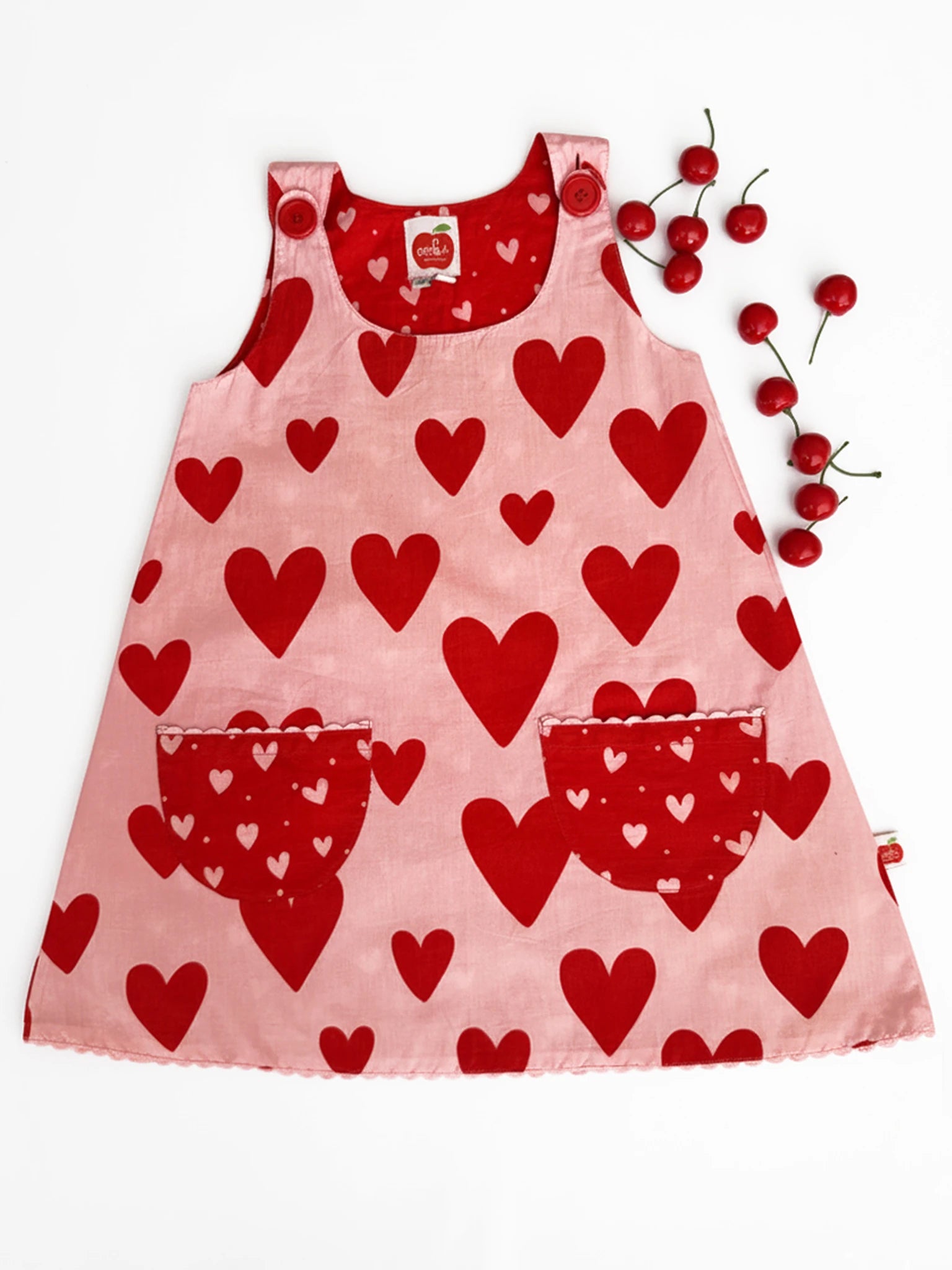 The Big Hearts Big Heart Gracie Reversible Dress is a pink cotton girls’ pinafore with large red heart patterns, two front pockets, and red strap buttons, shown with cherries on a white background.