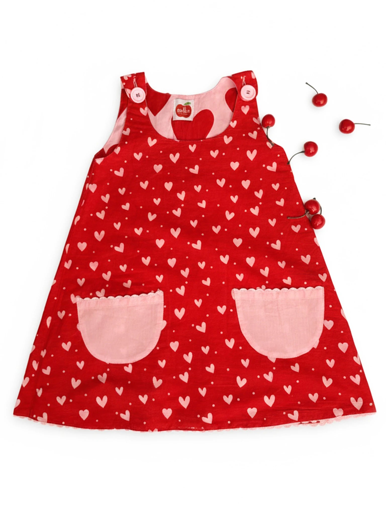 The Big Hearts Big Heart Gracie Reversible Dress, a red cotton girls' dress with white hearts and pink pockets, features two strap buttons. Cherries are placed beside the smock dress on a white background.