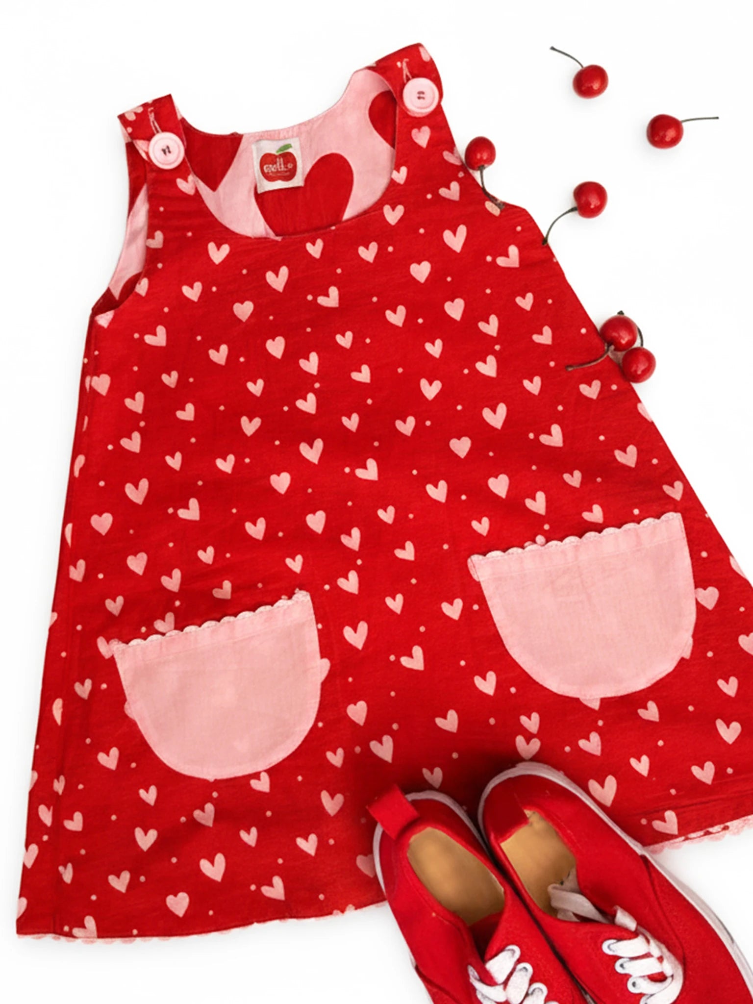 The Big Hearts Big Heart Gracie Reversible Dress features a red sleeveless cotton design with heart patterns, two pink pockets, red buttons, and comes with matching red sneakers, all set on a white background with scattered cherries.