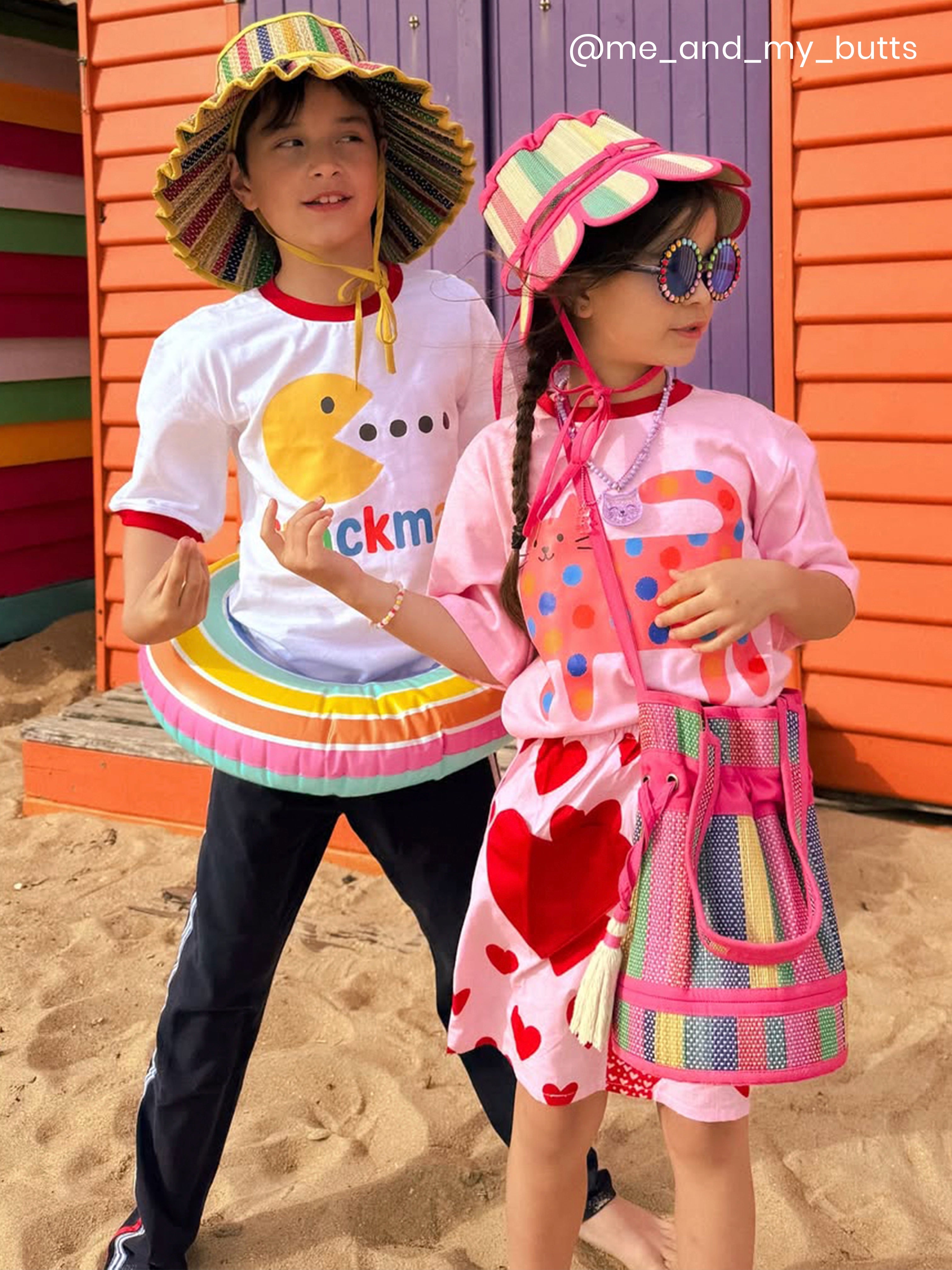 Two children in colorful beach attire standing on a sandy surface with a colorful wooden structure in the background.