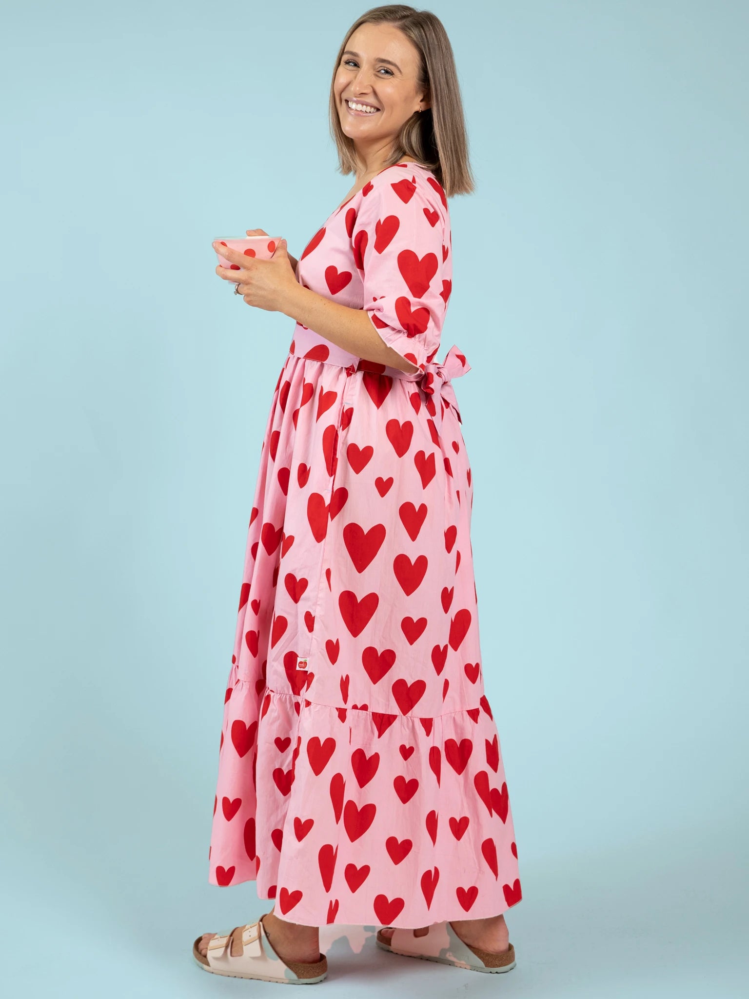 Woman smiles in the Big Hearts Women's Big Heart Maxi Dress—pink with red heart patterns—paired with white sandals and holding a mug against a light blue background, showcasing charming maternity style.