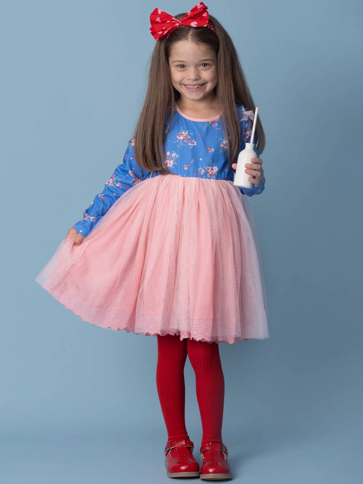 Young girl in a blue top and pink skirt with a red bow, holding a white object against a blue background