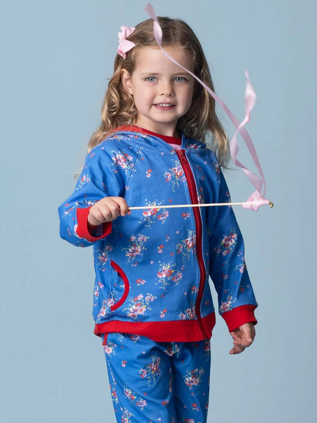 A young girl wears the Indigo Bloom Hoodie Jacket by Indigo Bloom with matching pants, holding a pink ribbon wand and standing against a plain light blue background.