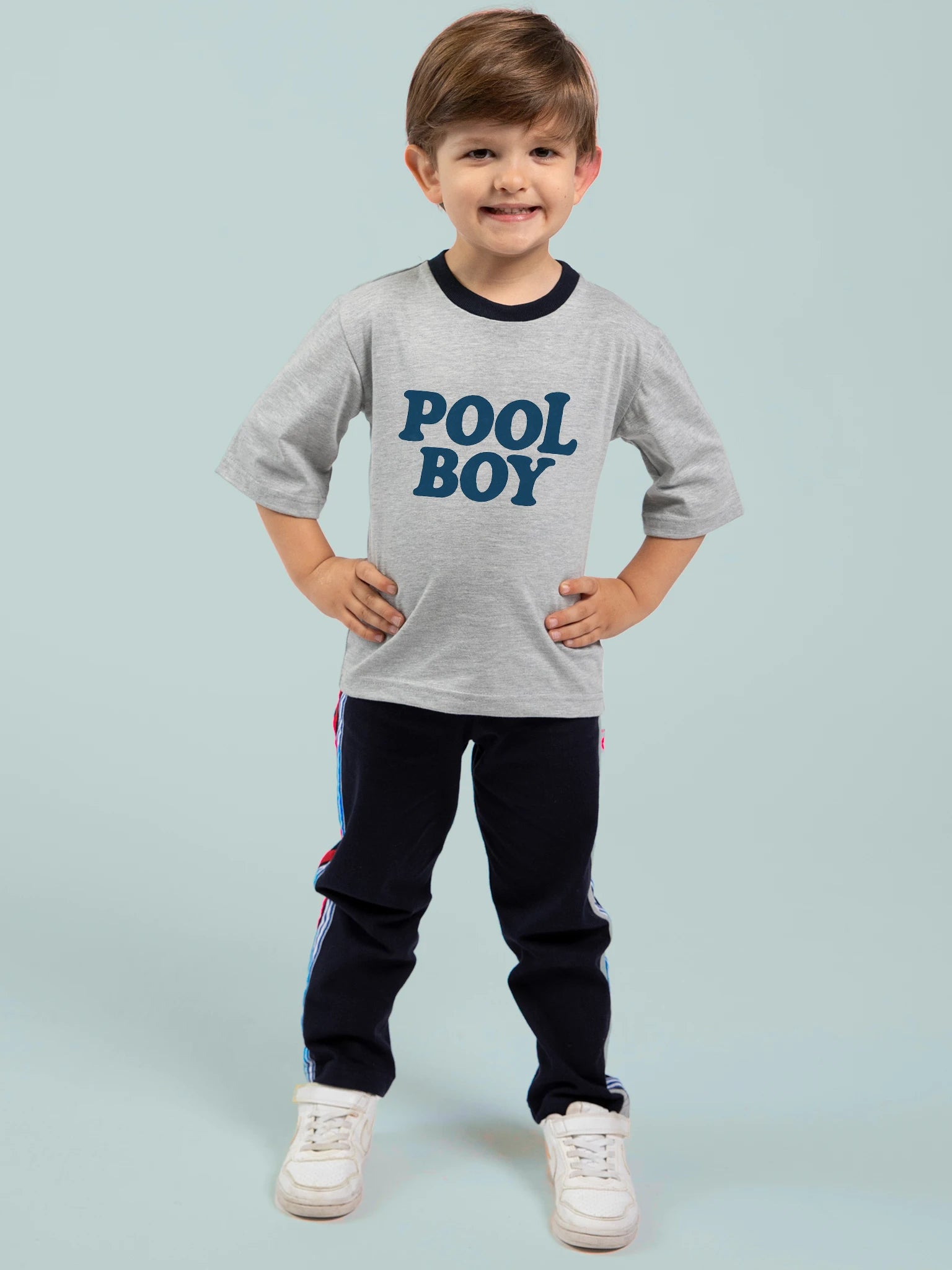 A smiling young boy stands with hands on hips in a grey Oobi Pool Boy Tee with navy binding—hand printed in Australia—paired with dark pants and white sneakers, set against a plain light blue background.