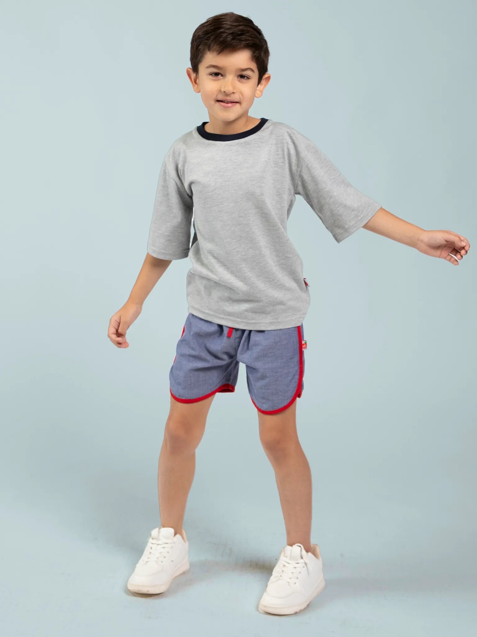 A boy stands on a light blue background wearing a grey t-shirt, white sneakers, and Essentials' Chambray Shorts With Retro Curve - FIND, crafted from 100% cotton with red trim.