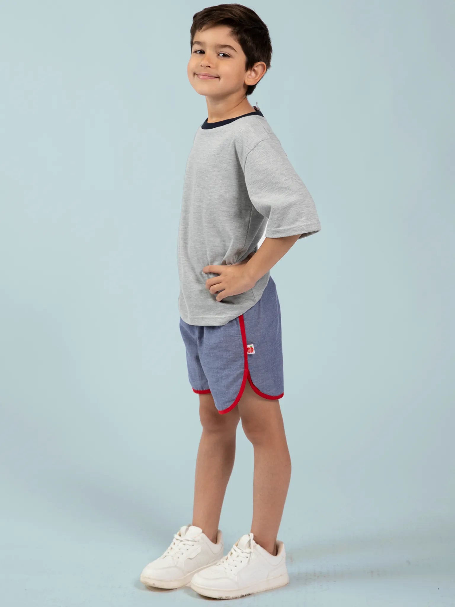 A boy stands sideways with hands on hips, wearing a grey 100% cotton T-shirt, Essentials Chambray Shorts With Retro Curve - FIND featuring red trim, and white sneakers against a plain light blue background.