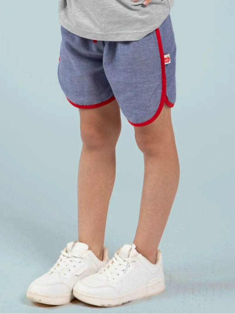 A child wears Essentials’ Chambray Shorts With Retro Curve - FIND, featuring red trim, and white sneakers, standing against a light blue background.