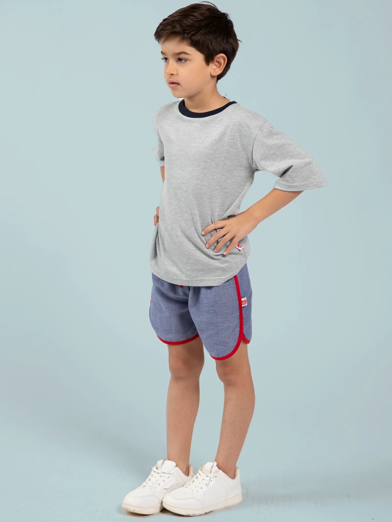 A young boy stands hands on hips, wearing a 100% cotton light grey tee, Essentials Chambray Shorts With Retro Curve by FIND featuring red trim, and white sneakers, set against a light blue background.
