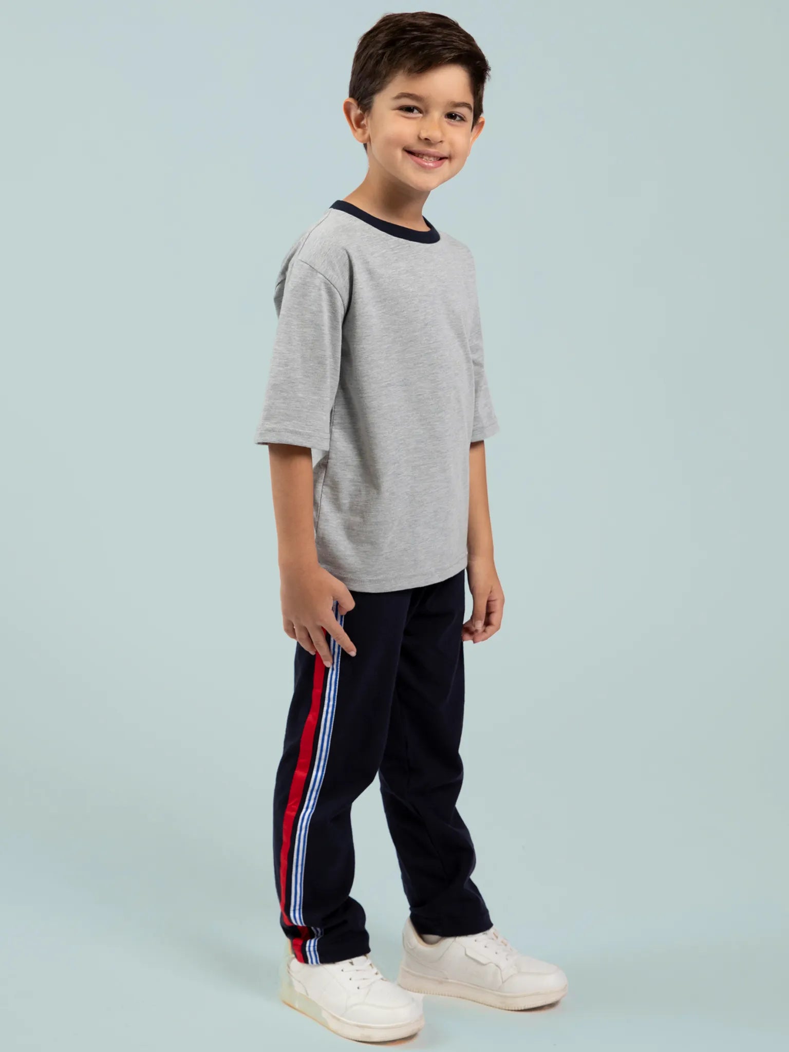 A young boy stands and smiles, wearing the Oobi Grey Marle Tee with Navy Binding 3078, navy track pants with red and blue side stripes, and white sneakers against a light gray background.