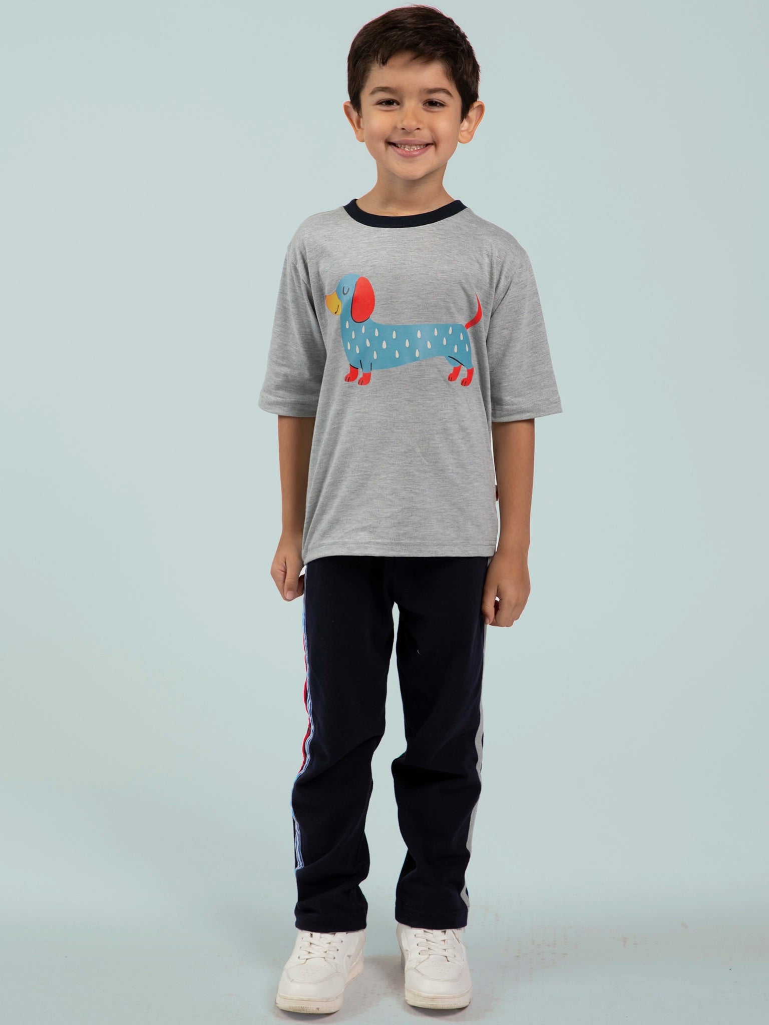 A young boy smiles in an Oobi Blue Sausage Dog Tee with Navy Binding, navy sweatpants, and white sneakers—showcasing stylish boys’ clothing against a plain light blue background.