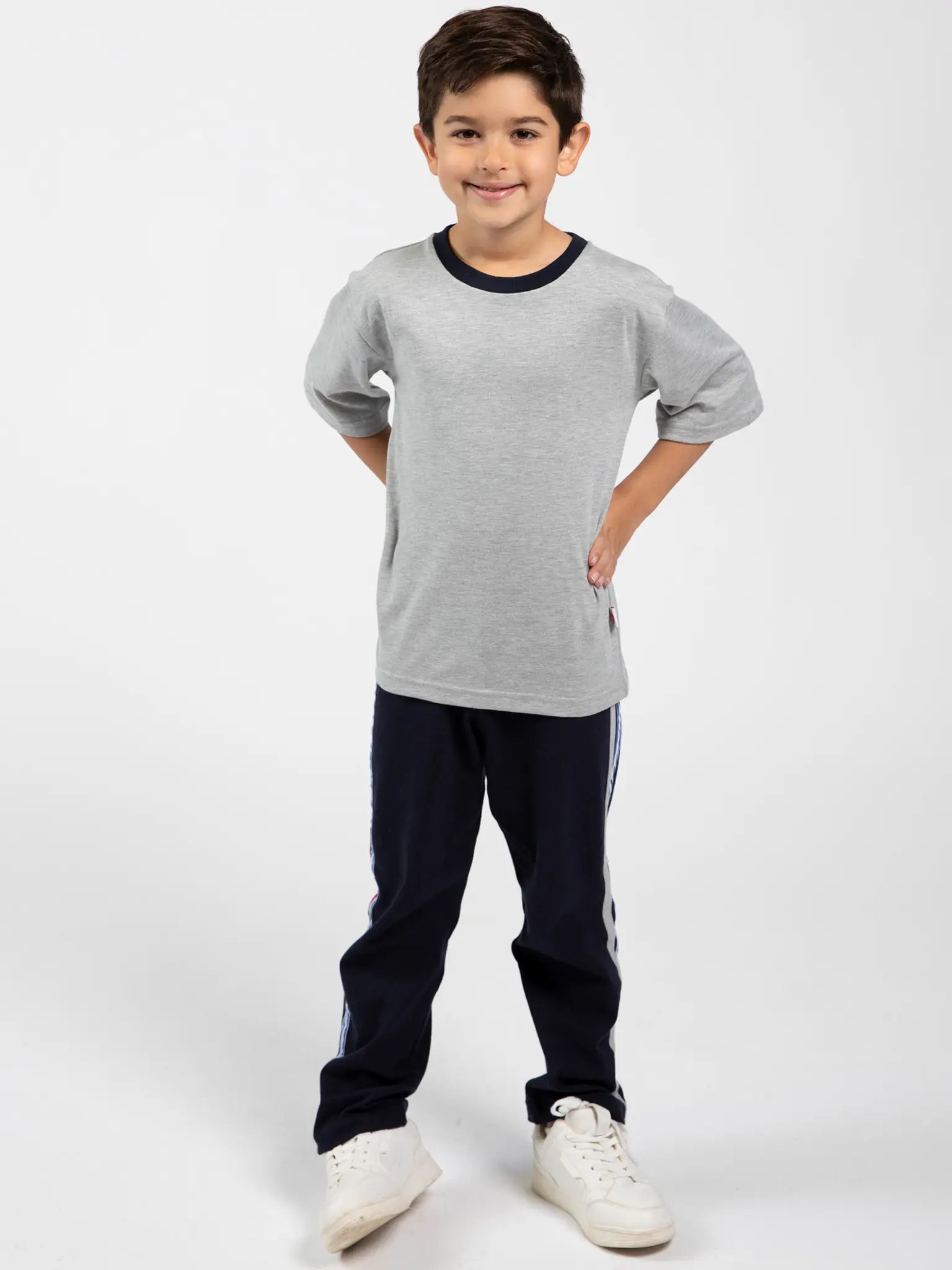 A young boy stands with hands on hips, wearing Oobi's Grey Marle Tee with Navy Binding 3078, navy striped sweatpants, and white sneakers against a plain white background.
