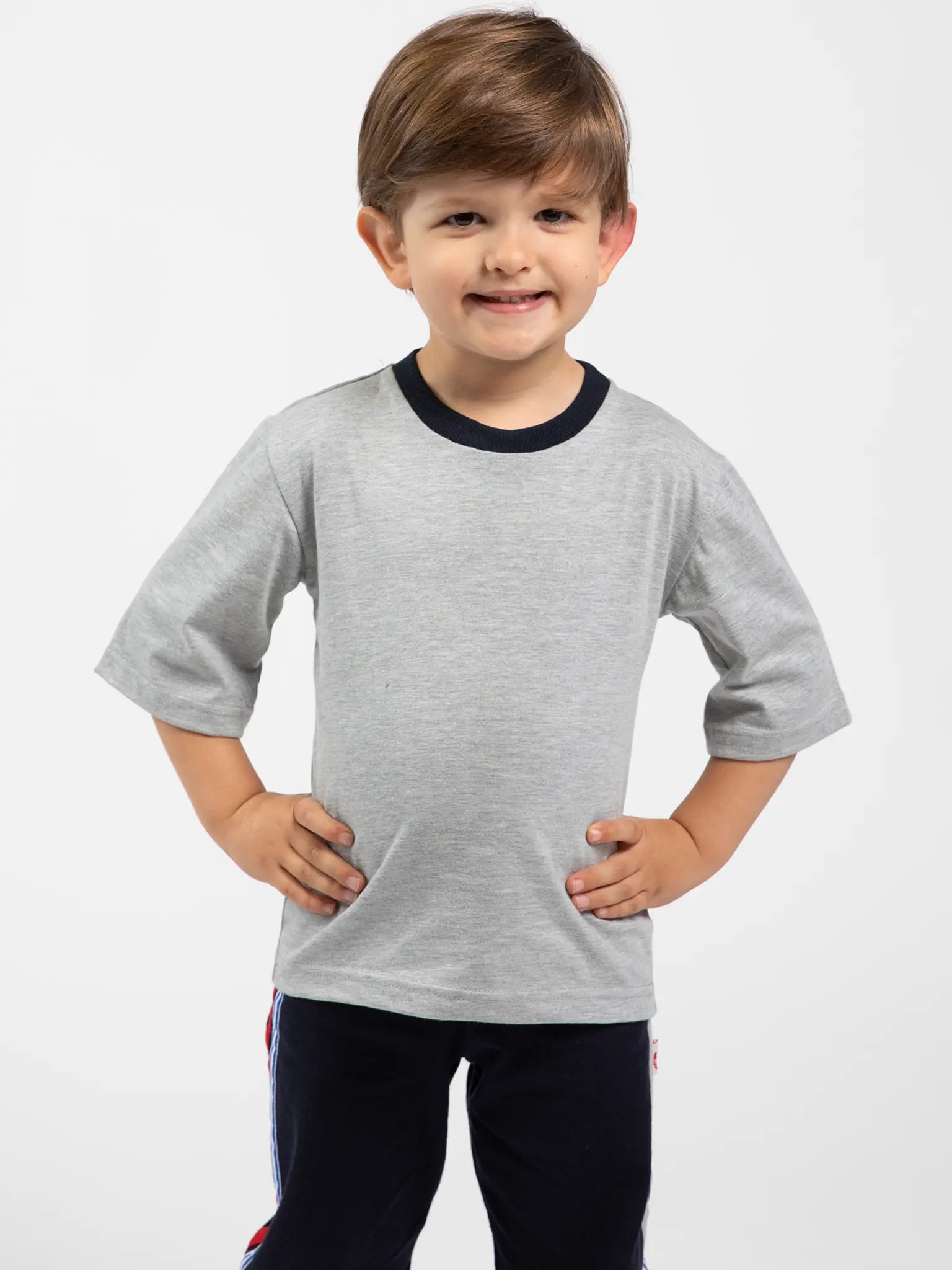 A young boy with light brown hair smiles, hands on hips, wearing the Oobi Grey Marle Tee with Navy Binding 3078 and dark pants against a plain white background.