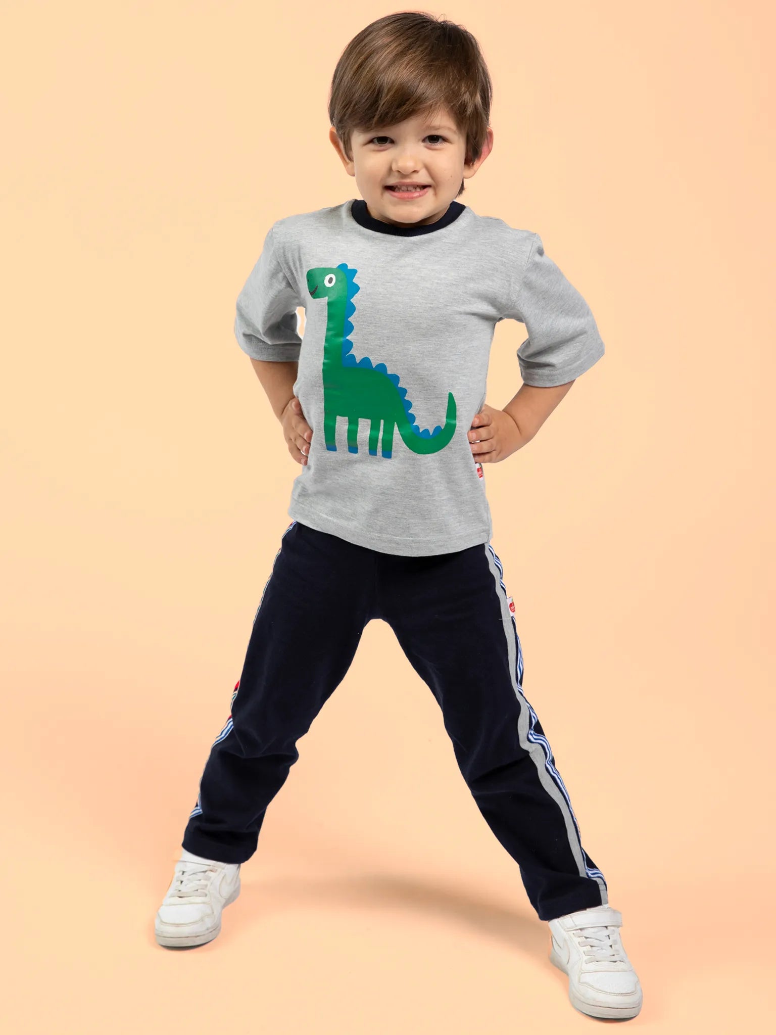 A young child stands confidently with hands on hips, wearing Oobi's Dino-mite Tee with Navy Binding—made from 100% cotton and featuring a green dinosaur graphic—paired with navy blue pants with white stripes, against a plain peach background.