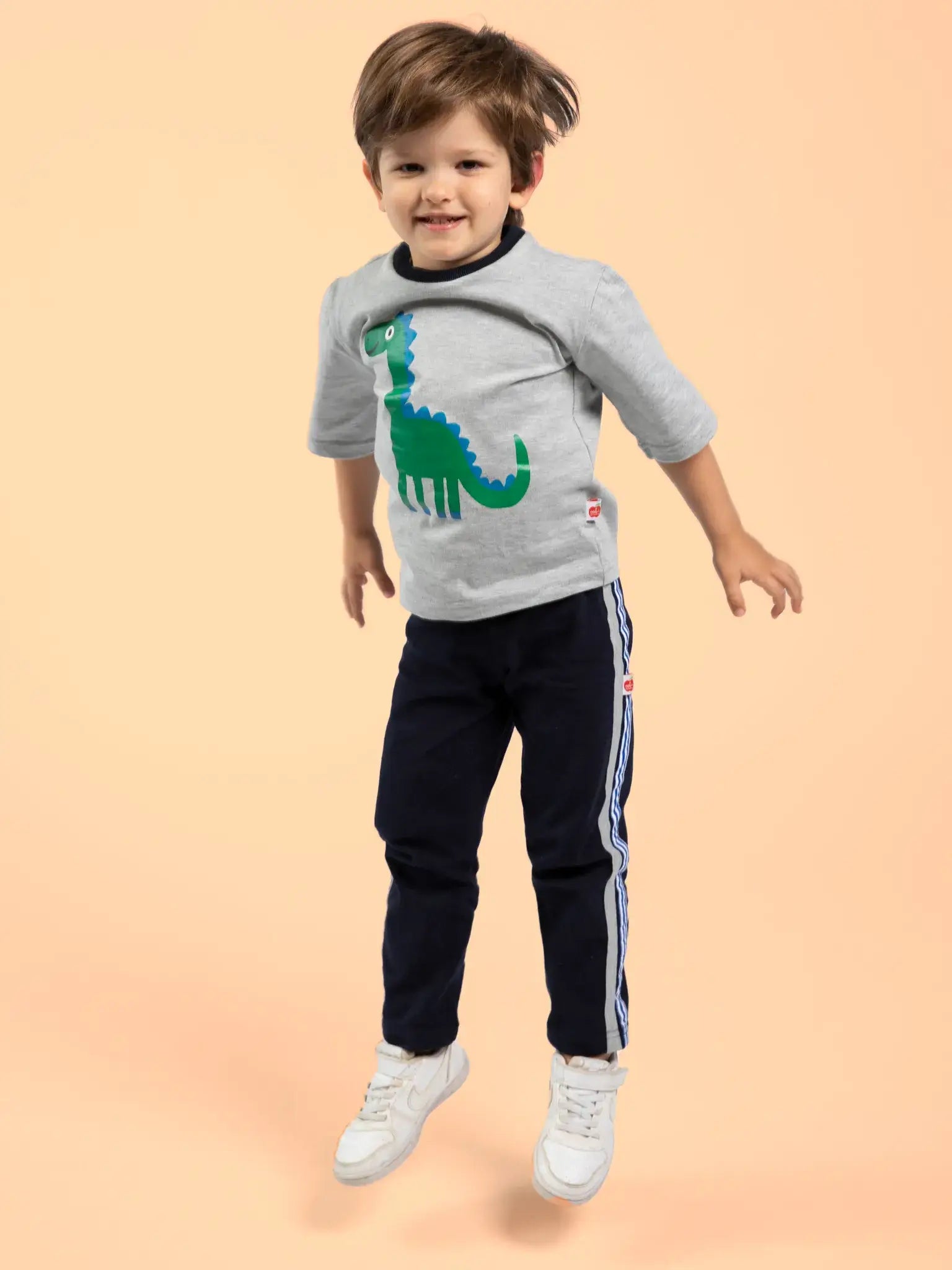 A young child in Oobi’s Dino-mite Tee with Navy Binding, navy track pants, and white sneakers jumps against a plain peach background.