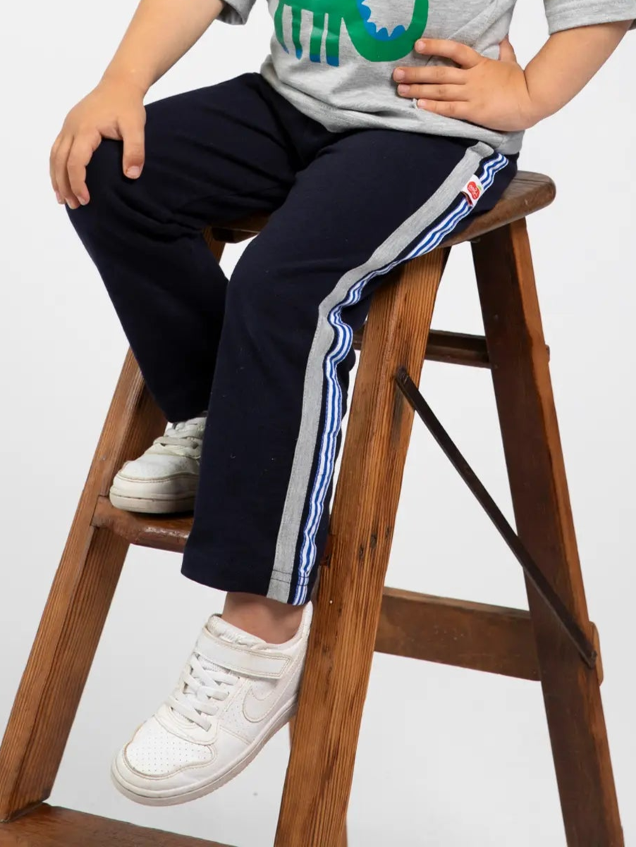 A child in a grey t-shirt, Essentials Navy Jersey 2 Stripe Pants, and white sneakers sits on a wooden step ladder.