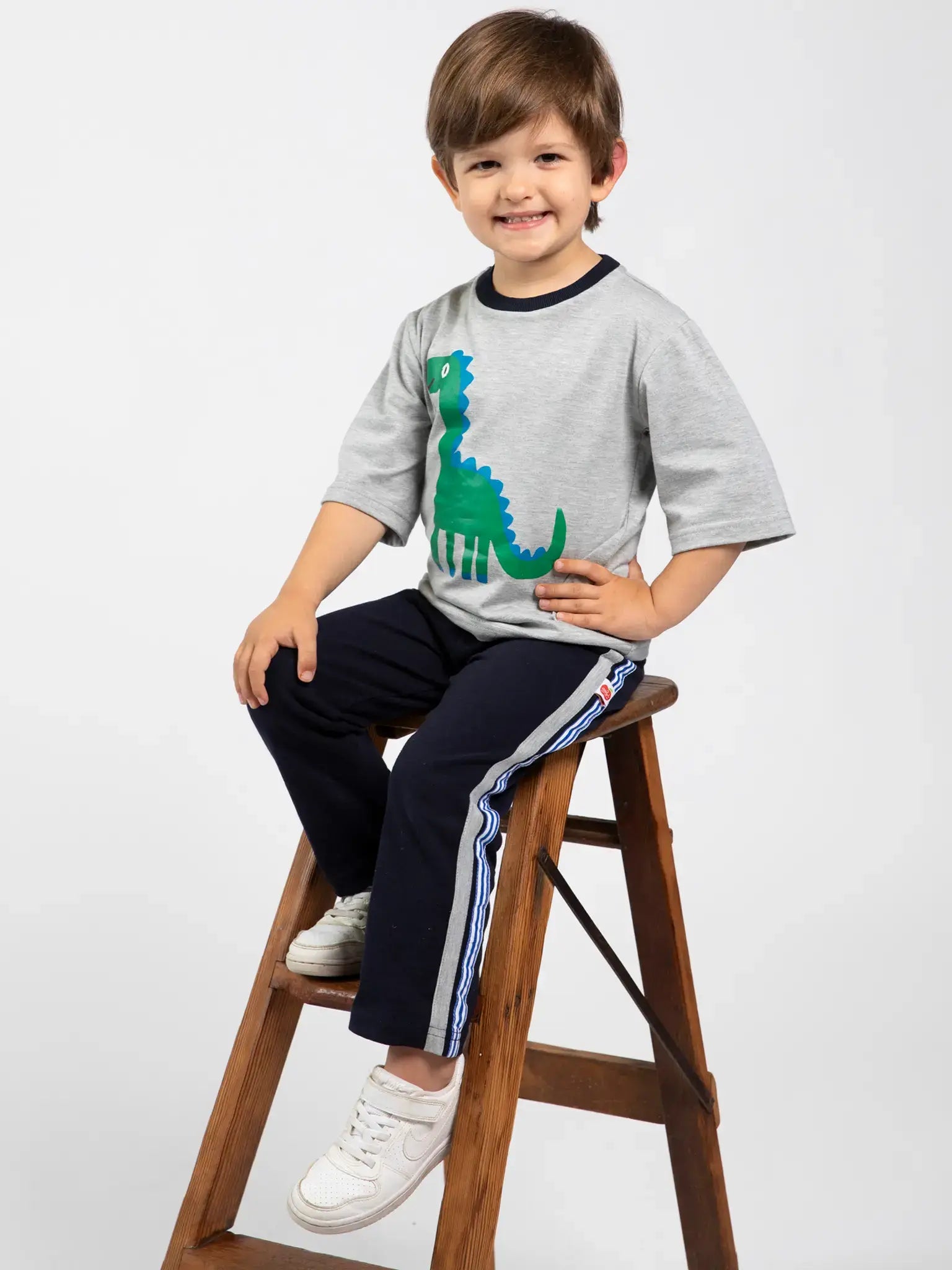A young boy smiles on a wooden ladder, wearing Oobi’s Dino-mite Tee with Navy Binding—100% cotton, hand printed in Australia—paired with navy sweatpants and white sneakers.
