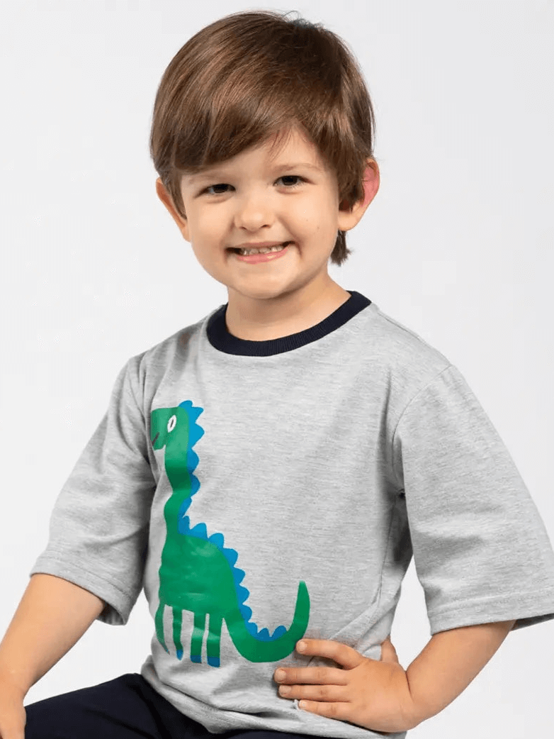 A young boy with brown hair smiles in the Oobi Dino-mite Tee with Navy Binding—a 100% cotton grey tee featuring a green and blue dinosaur graphic. Ideal for boys tees collections, shown against a plain light background.