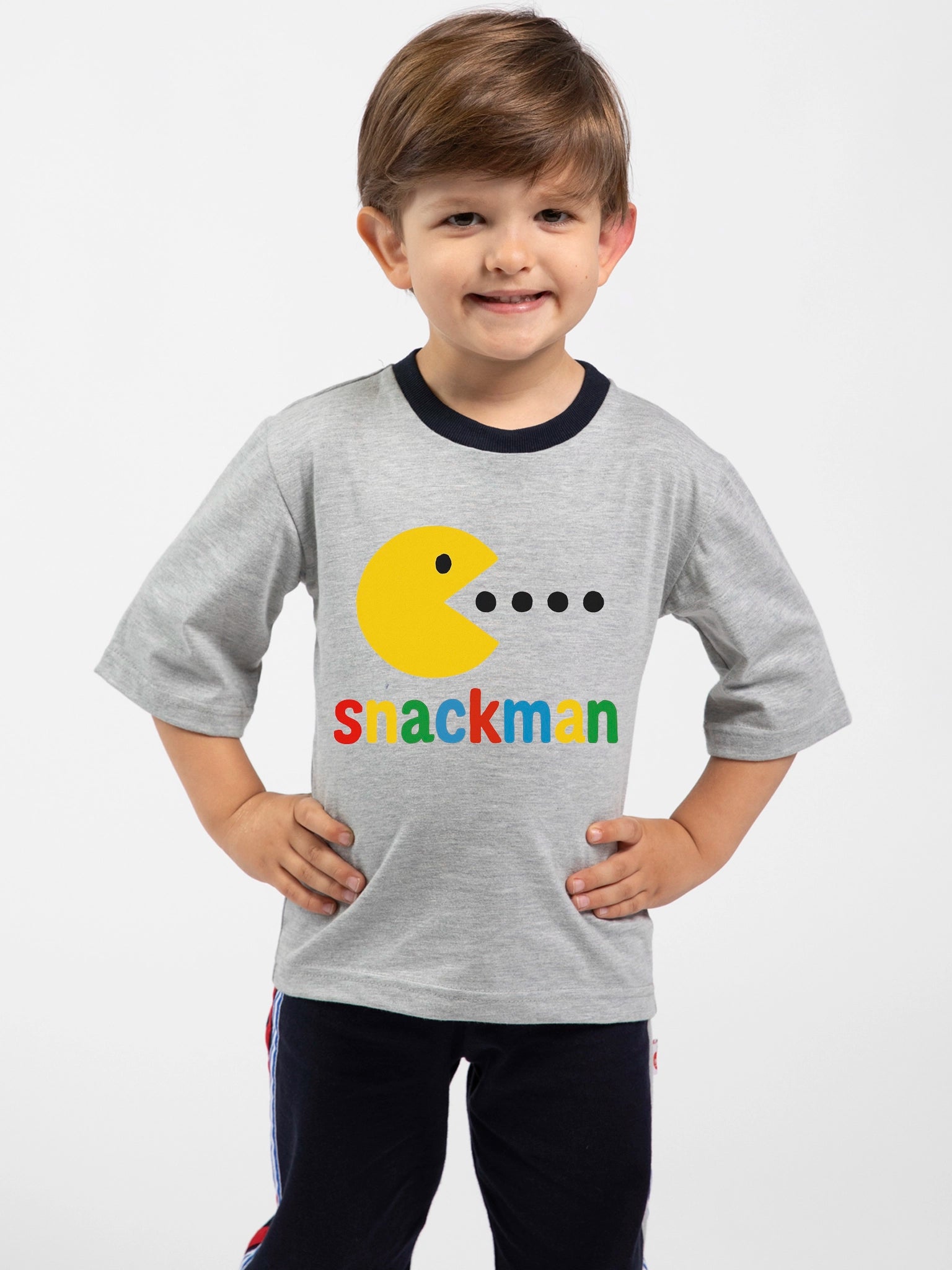 A young boy smiles with hands on hips, wearing the Essentials Snackman Printed Tee—an oversized gray shirt with a yellow Pac-Man-style graphic and “snackman” in colorful letters. Hand printed in Australia. Free personalisation available!.