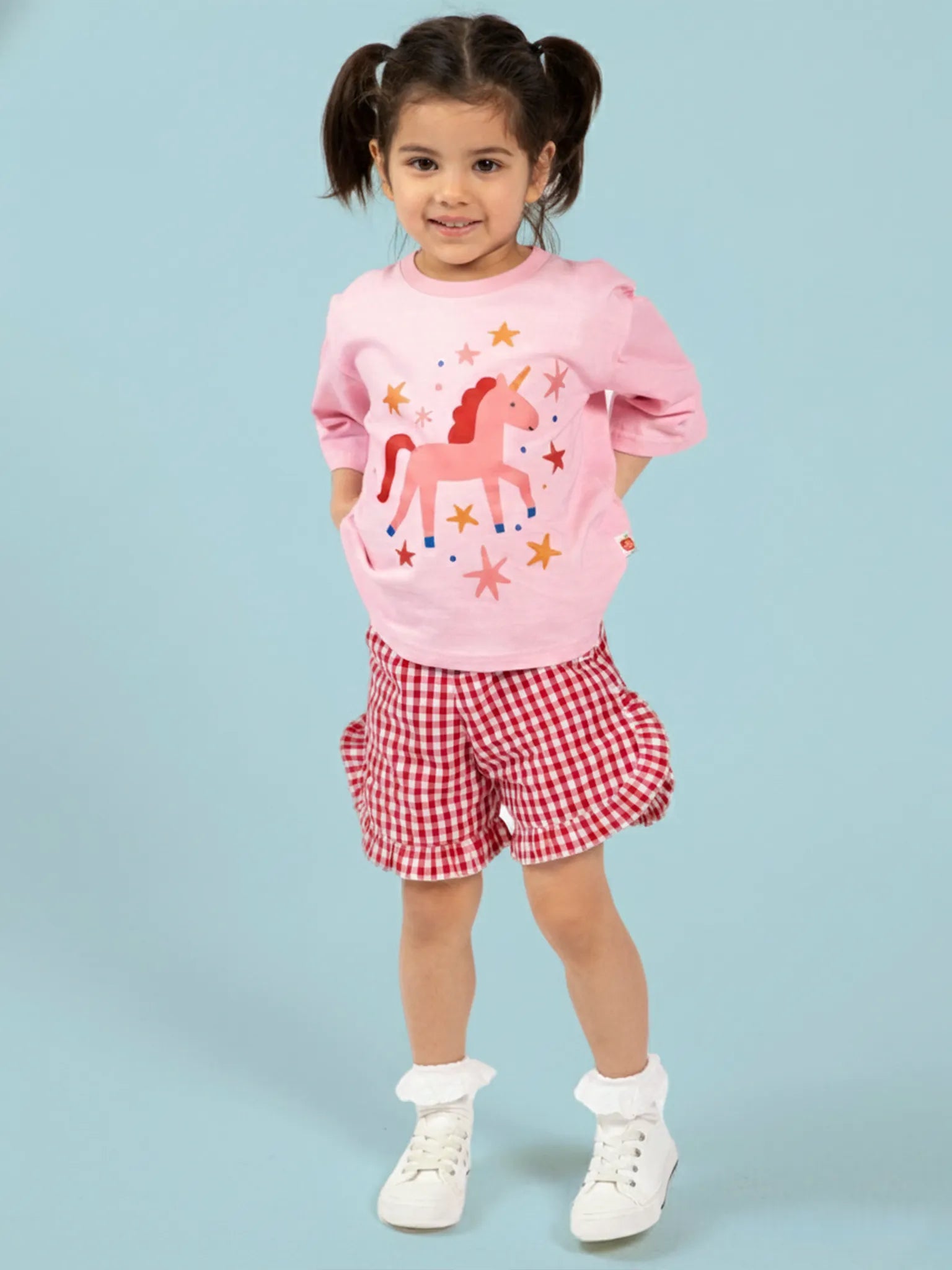 A young girl with pigtails stands against a light blue background, wearing Red Gingham Ruffle Shorts by Red Gingham and a pink unicorn shirt.