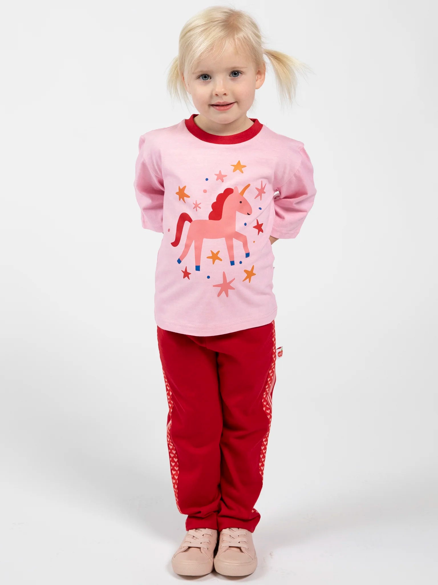 A young child with blonde pigtails stands facing forward, wearing a pink unicorn shirt, Essentials Red Jersey 2 Stripe Pants made from 100% cotton, and pink shoes, against a plain white background.