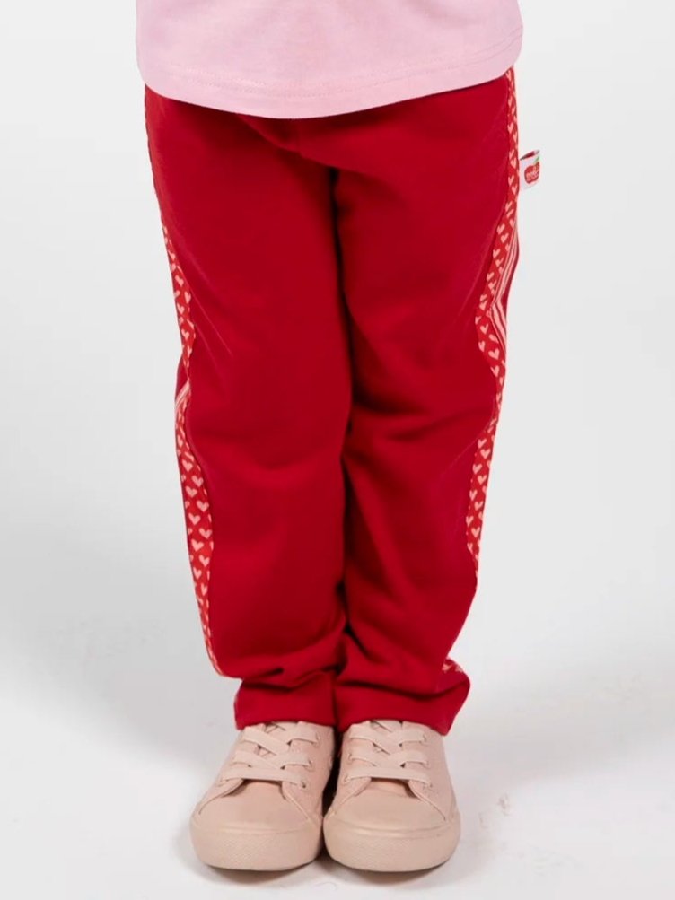 A child in Essentials Red Jersey 2 Stripe Pants and light pink sneakers stands against a plain background—an on-trend girls’ sportswear look.