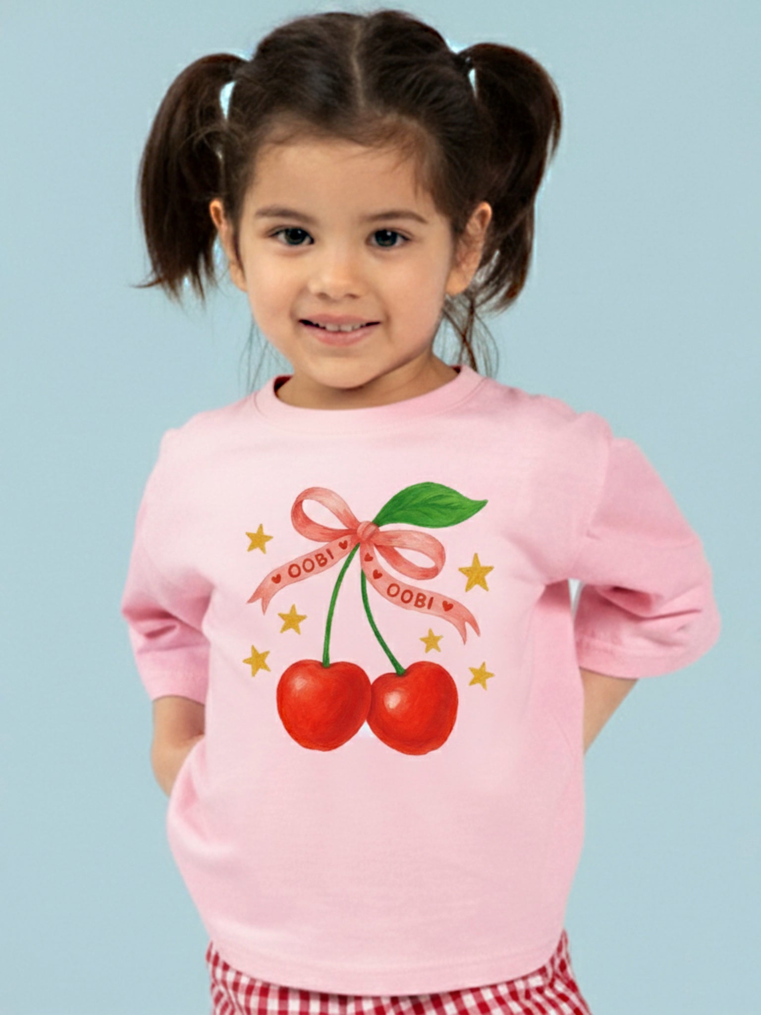 A young girl with pigtails wears the Essentials Pink Cherry Tee with Red Binding, which features two illustrated cherries tied with a pink ribbon and surrounded by yellow stars.