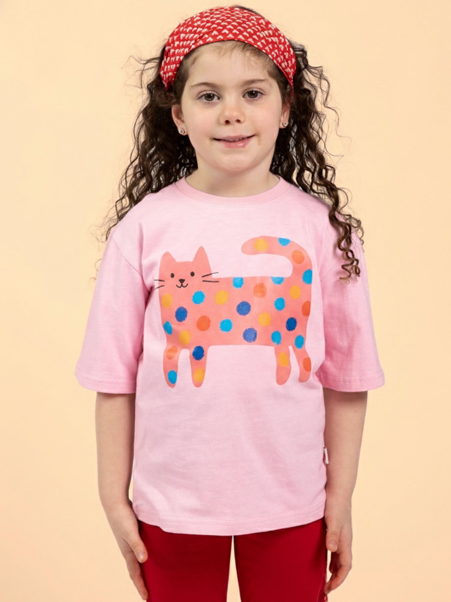 Young girl with long curly hair and a red headband wears the Essentials Quirky Cat Pink Tee with Red Binding and red pants, standing against a plain light background.
