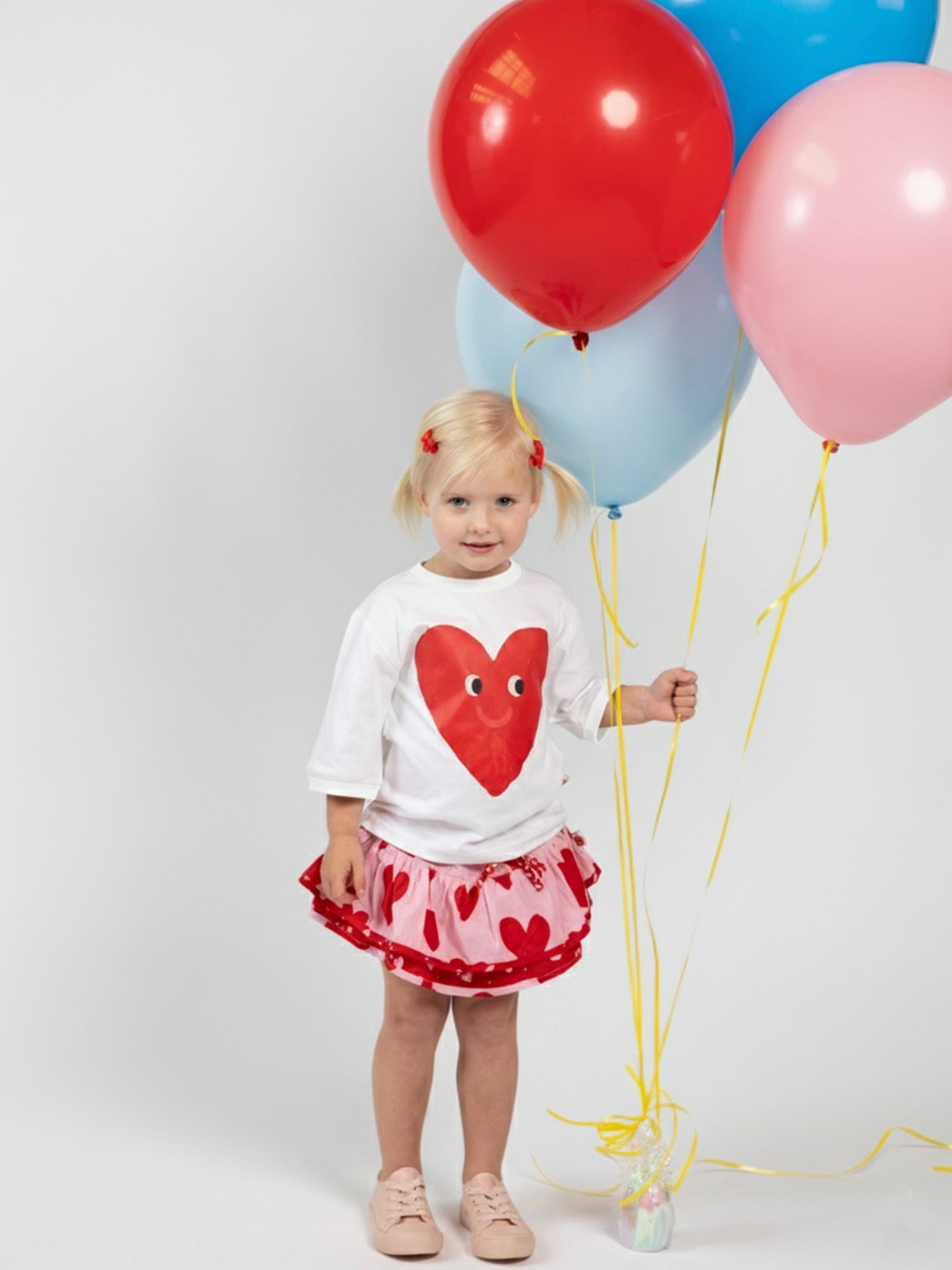 A young girl holds three balloons, wearing the Essentials Happy Heart Tee with Red Binding, an oversized heart-patterned skirt, and pink shoes, set against a plain light background.