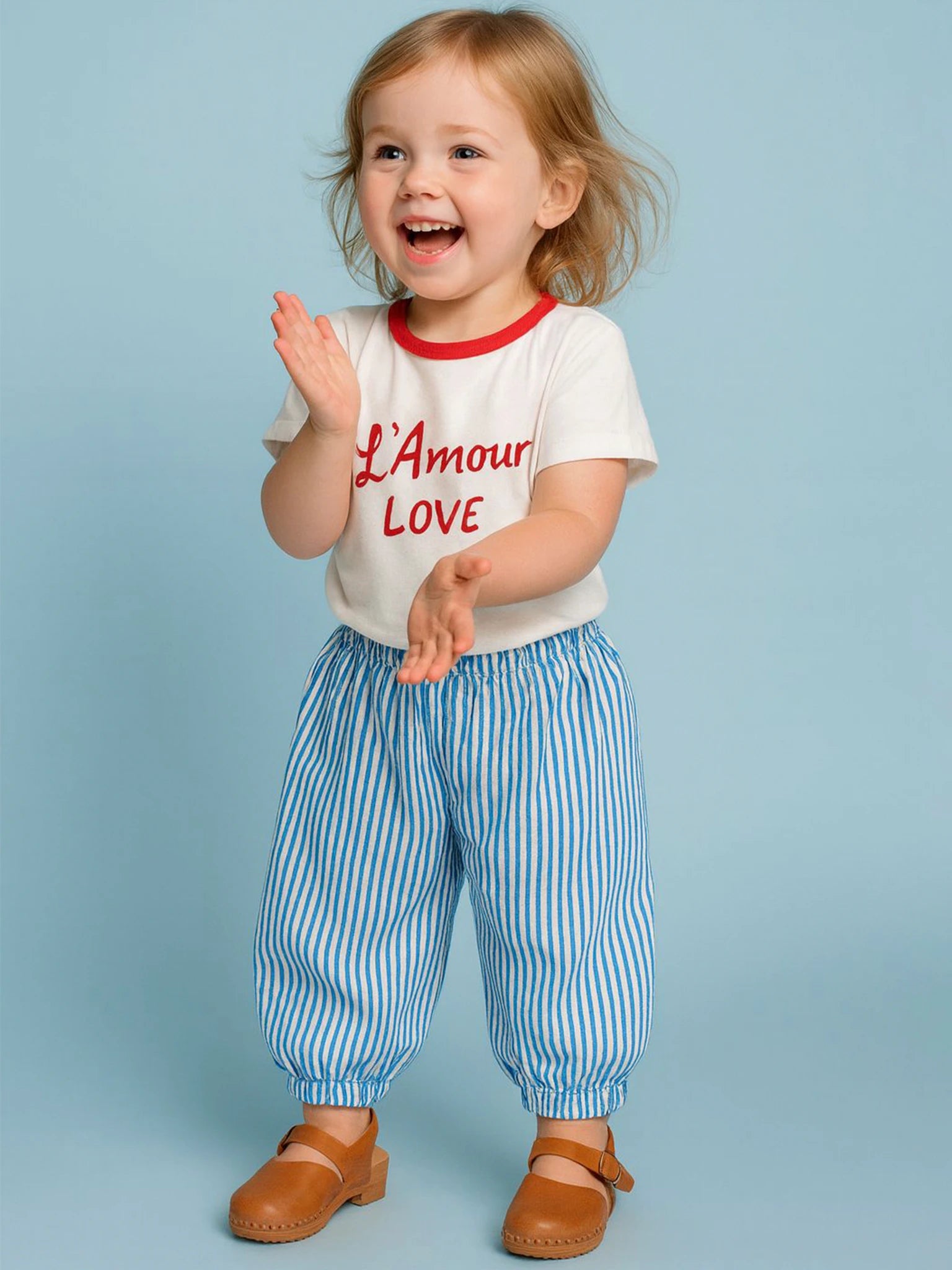 A young child smiles and claps in an oversized 100% cotton Essentials L'Amour (Love) Tee with red binding, paired with blue striped pants and brown shoes, standing against a light blue background.
