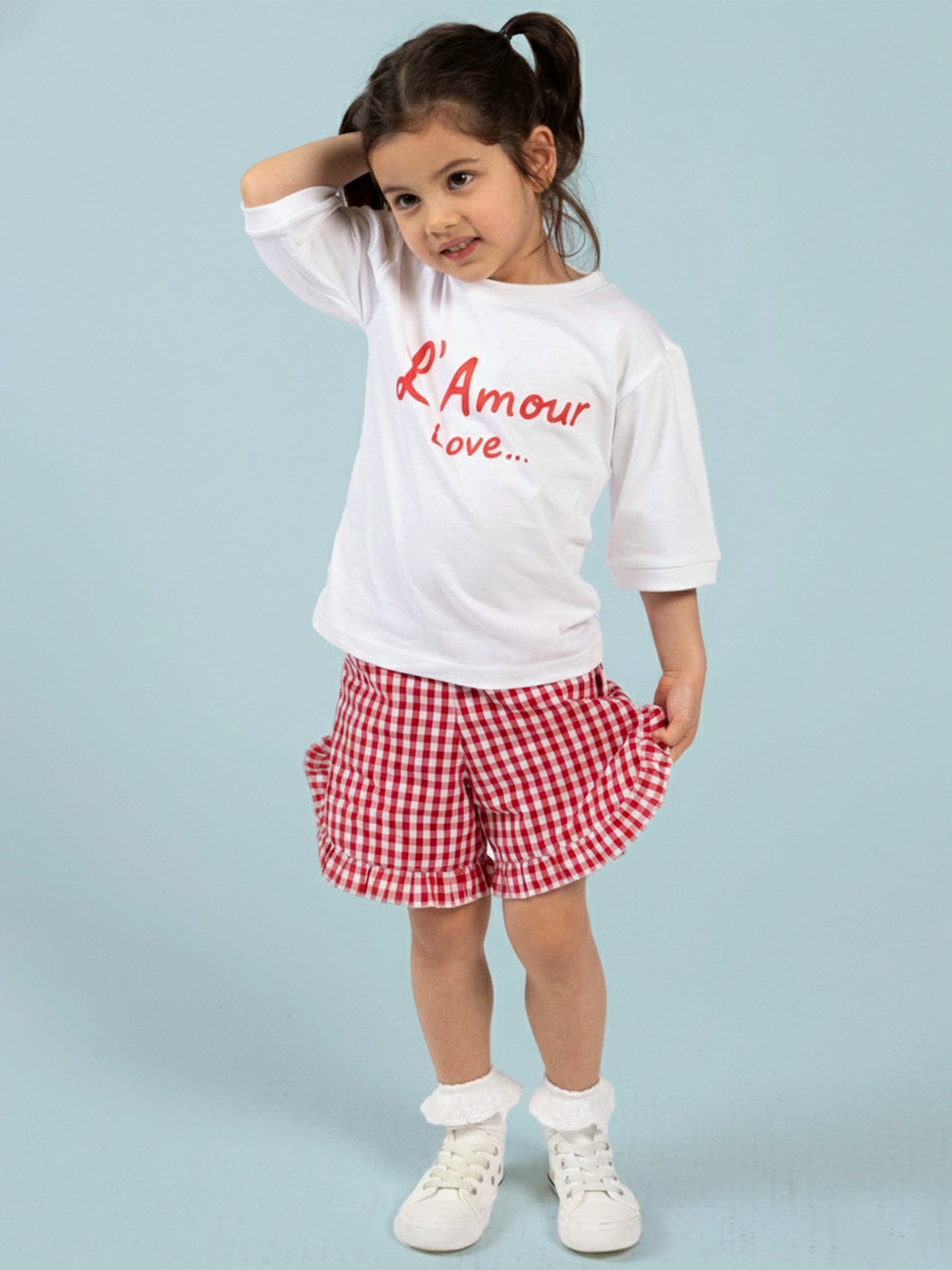 A young girl wears Red Gingham brand ruffle shorts with a red gingham pattern and an elastic waist, paired with a white T-shirt that has "L'Amour Love..." in red, standing against a light blue background.