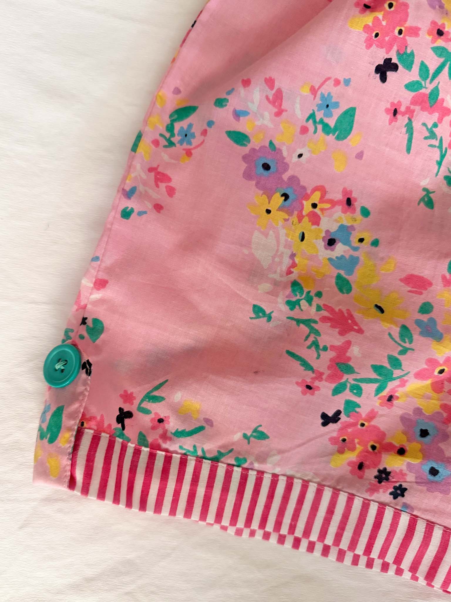 A close-up of the Confetti Floral Josie Playsuit by Confetti Floral shows pink cotton fabric with a colorful floral pattern, blue button, and pink-and-white striped trim, displayed on a white surface.