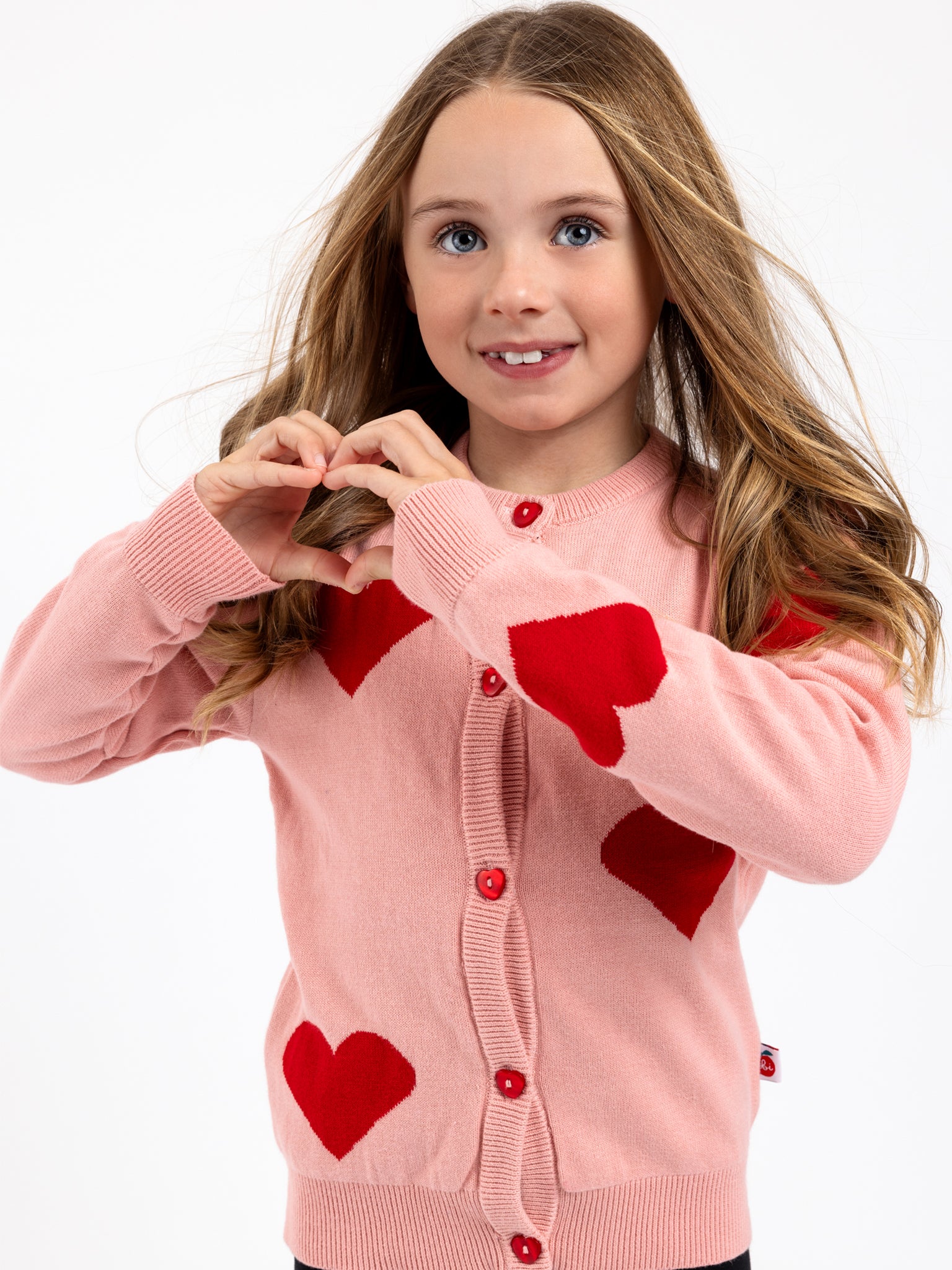 A young girl with long hair, dressed in Oobi’s Love Forever Outfit with FREE HAT!, smiles at the camera and forms a heart shape with her hands.
