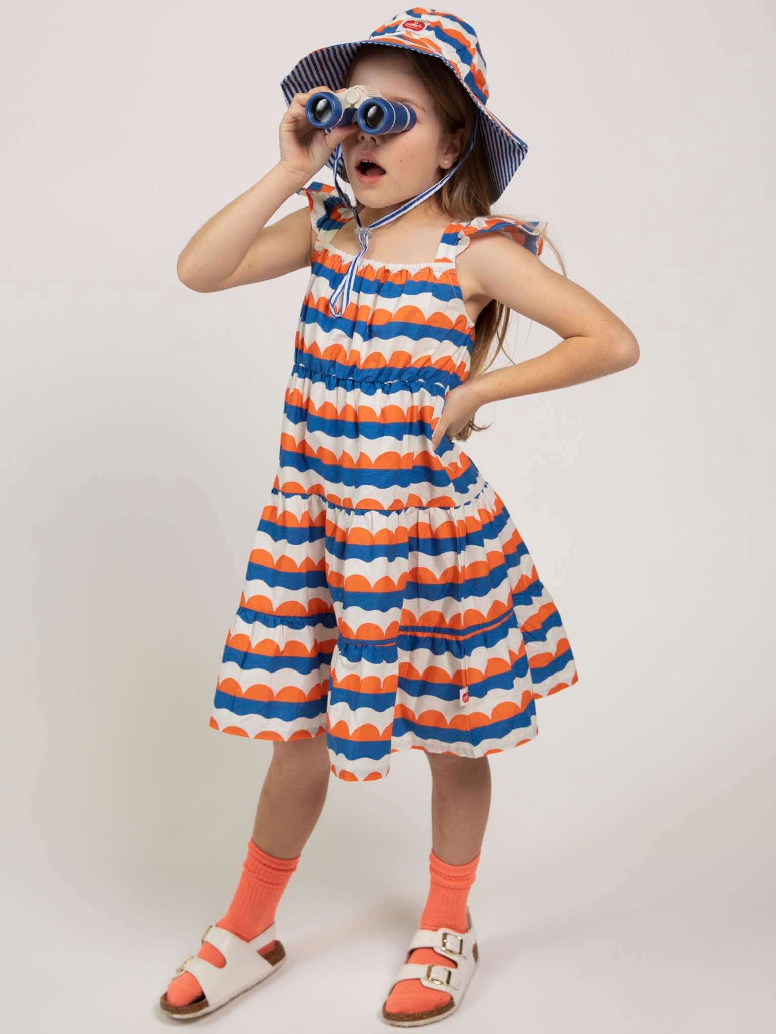 A young girl wears the Nautical Holiday Dress by Nautical, featuring colorful stripes and a matching hat, as she looks through blue binoculars with one hand in her pocket, set against a plain background.