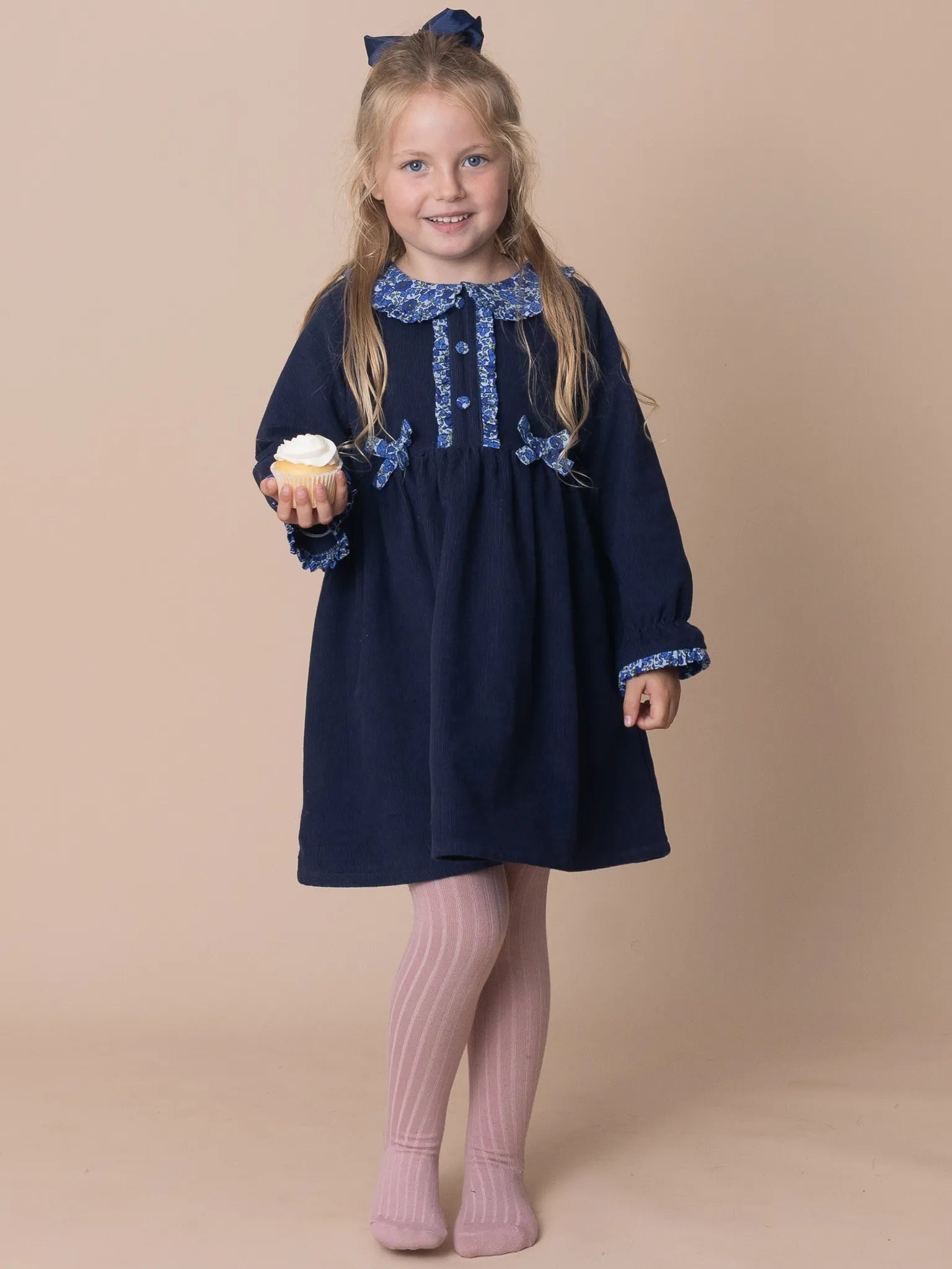 Young girl wearing a navy dress with lace details, holding a cupcake against a beige background