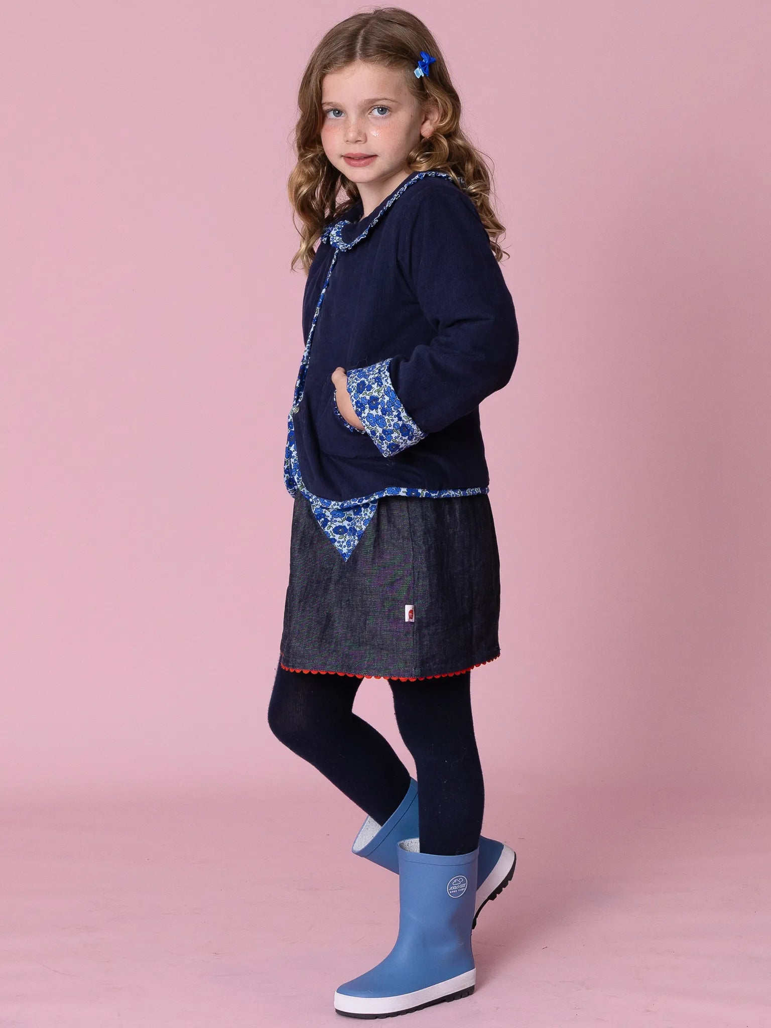 Young girl wearing a navy dress with floral patterns and blue boots on a pink background