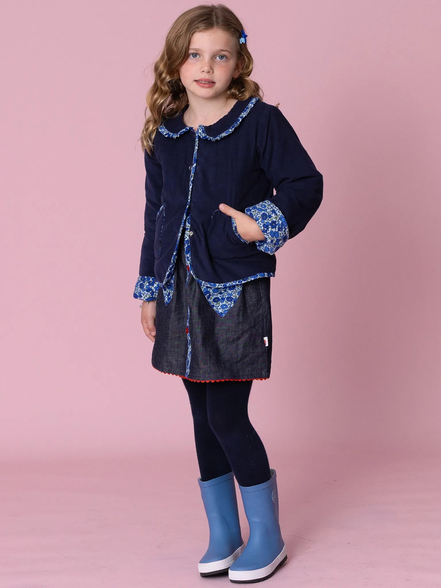 Young girl wearing a navy dress with blue accents and blue boots on a pink background