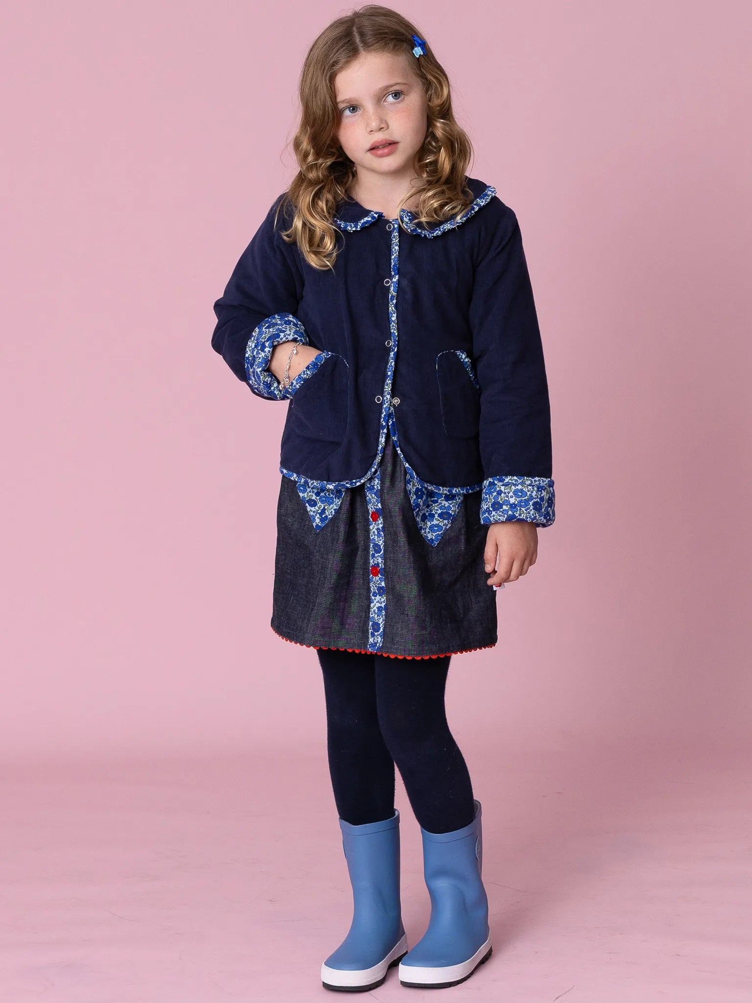 Young girl wearing a navy coat with floral patterns, black leggings, and blue boots on a pink background