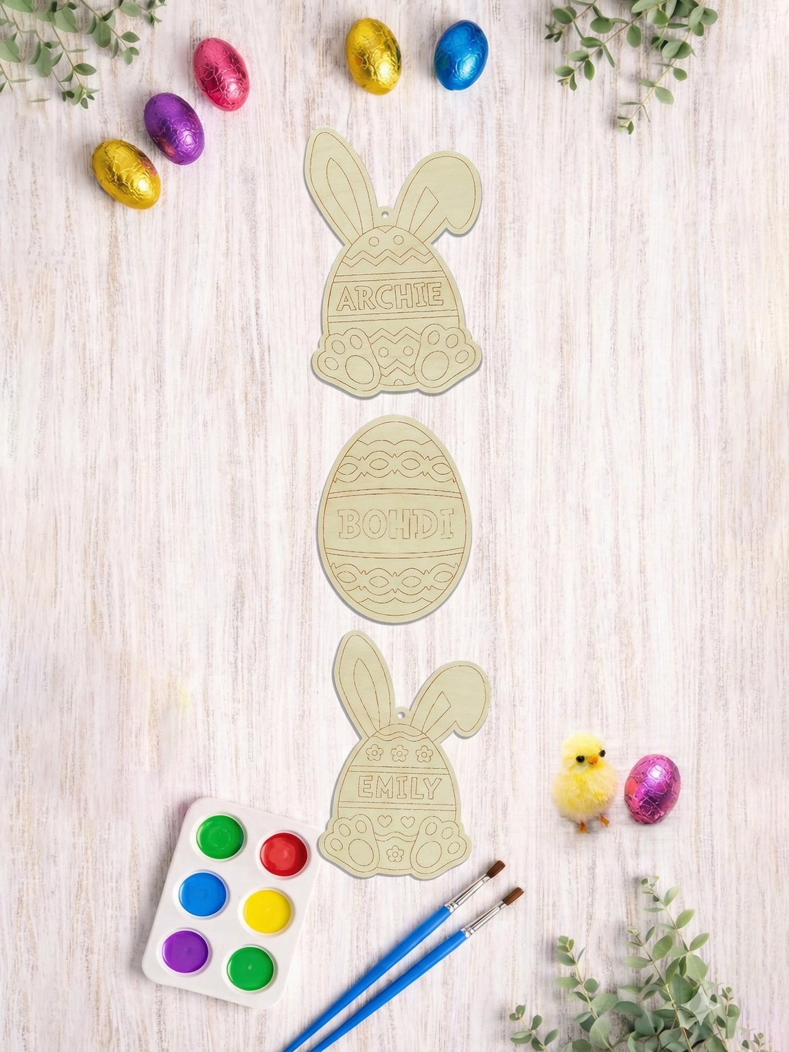 Easter-themed cookie cutters with 'ARCHIE' and 'BOBBY' on a wooden surface with colorful Easter eggs and paint supplies.