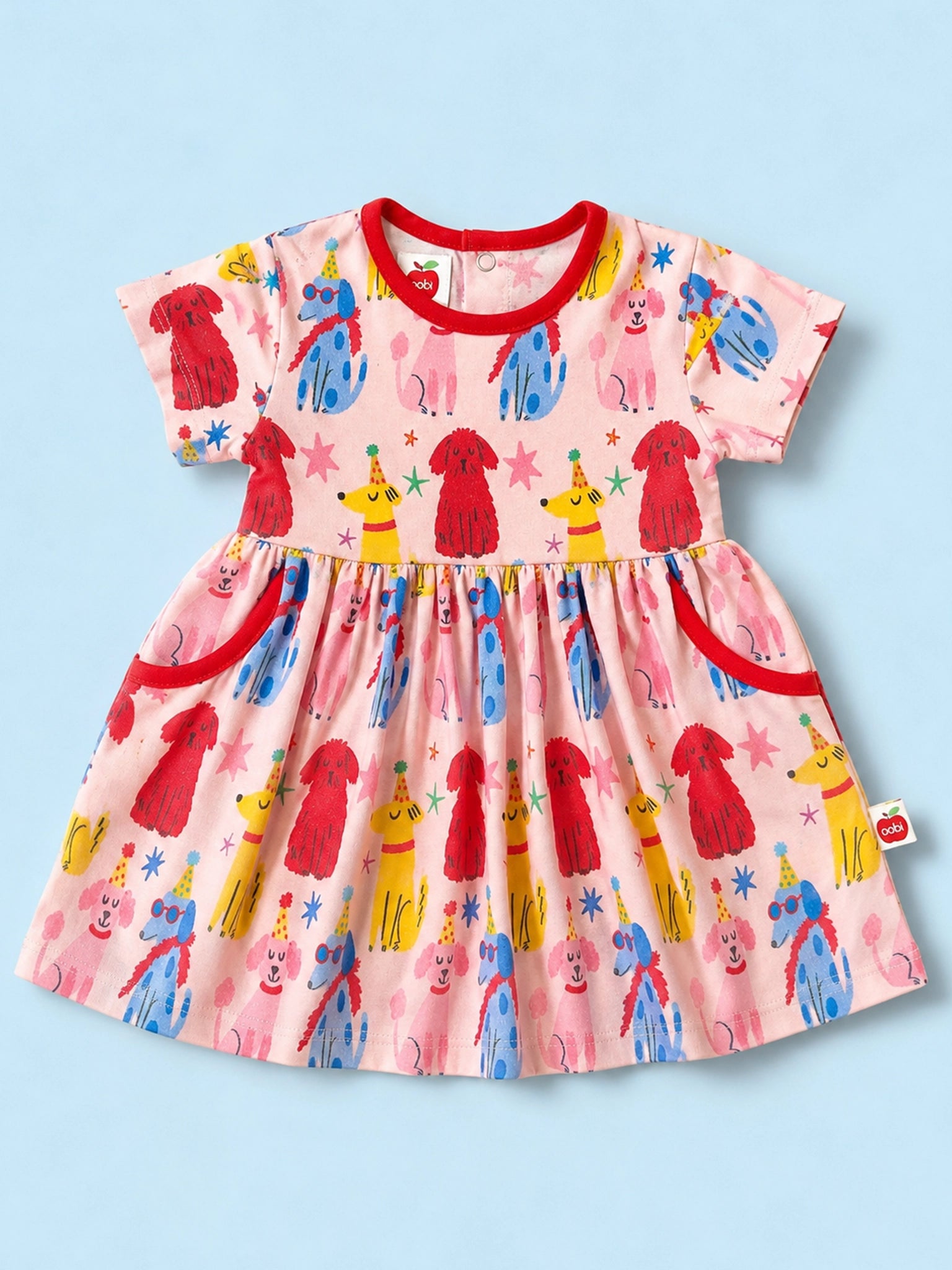 A short-sleeve baby dress with a pink base and a colorful print of cartoon dogs, stars, and faces, featuring red trim on the collar, cuffs, and pockets, laid flat on a light blue background.