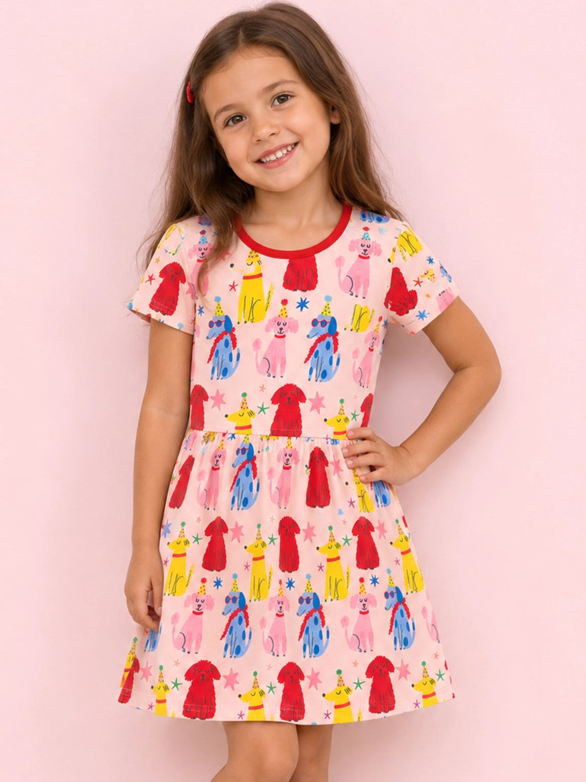 A young girl with long brown hair stands against a solid pink background wearing a short-sleeve pink dress with a colorful cartoon pattern of figures in red, blue, yellow, and pink. She is wearing red Mary Jane shoes.