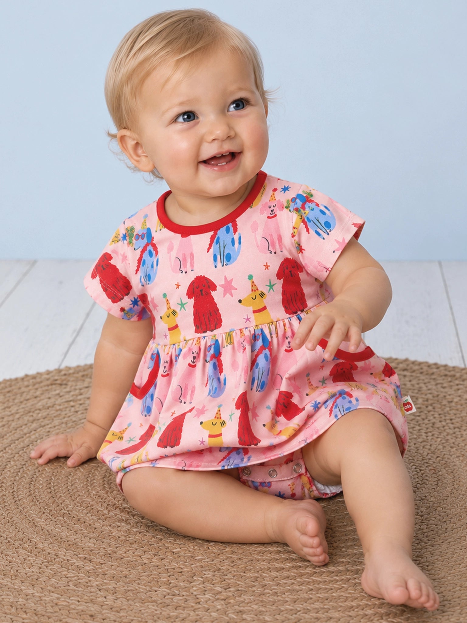 A smiling baby wearing a short-sleeve dress with a pink base and a colorful print of cartoon dogs, stars, and faces, featuring red trim on the collar, cuffs, and pockets on a light blue background.