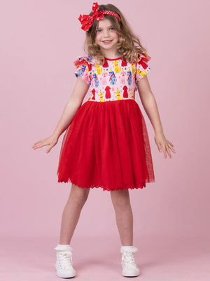 A young girl modeling a red and white dress with a colorful patterned bodice, white tulle skirt, and a red bow headband, standing against a solid pink background.