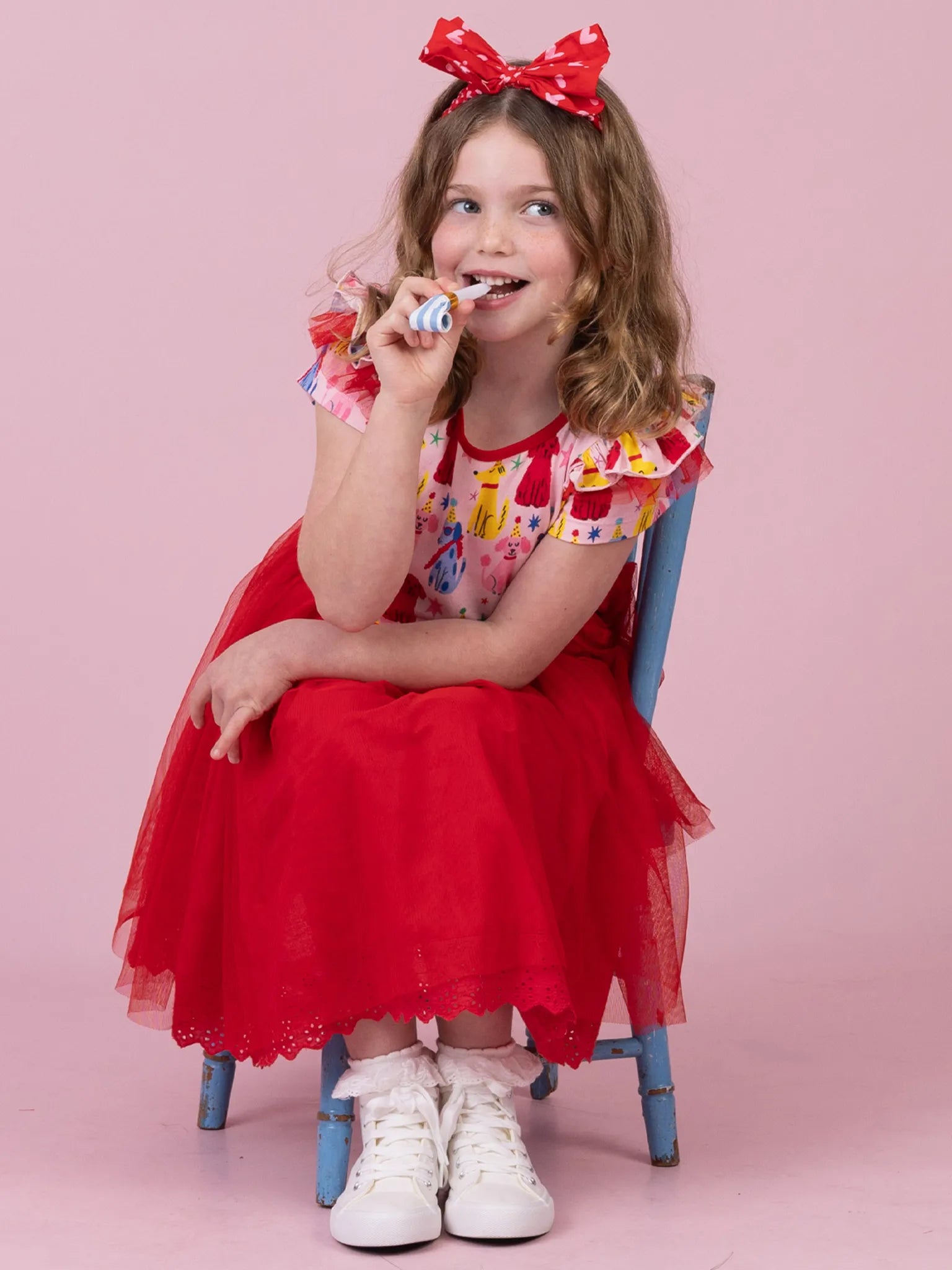 A young girl with a red polka-dot bow sits on a blue chair against a solid pink background, wearing a red tulle skirt, a colorful patterned top, and white sneakers while holding a small toy to her mouth.