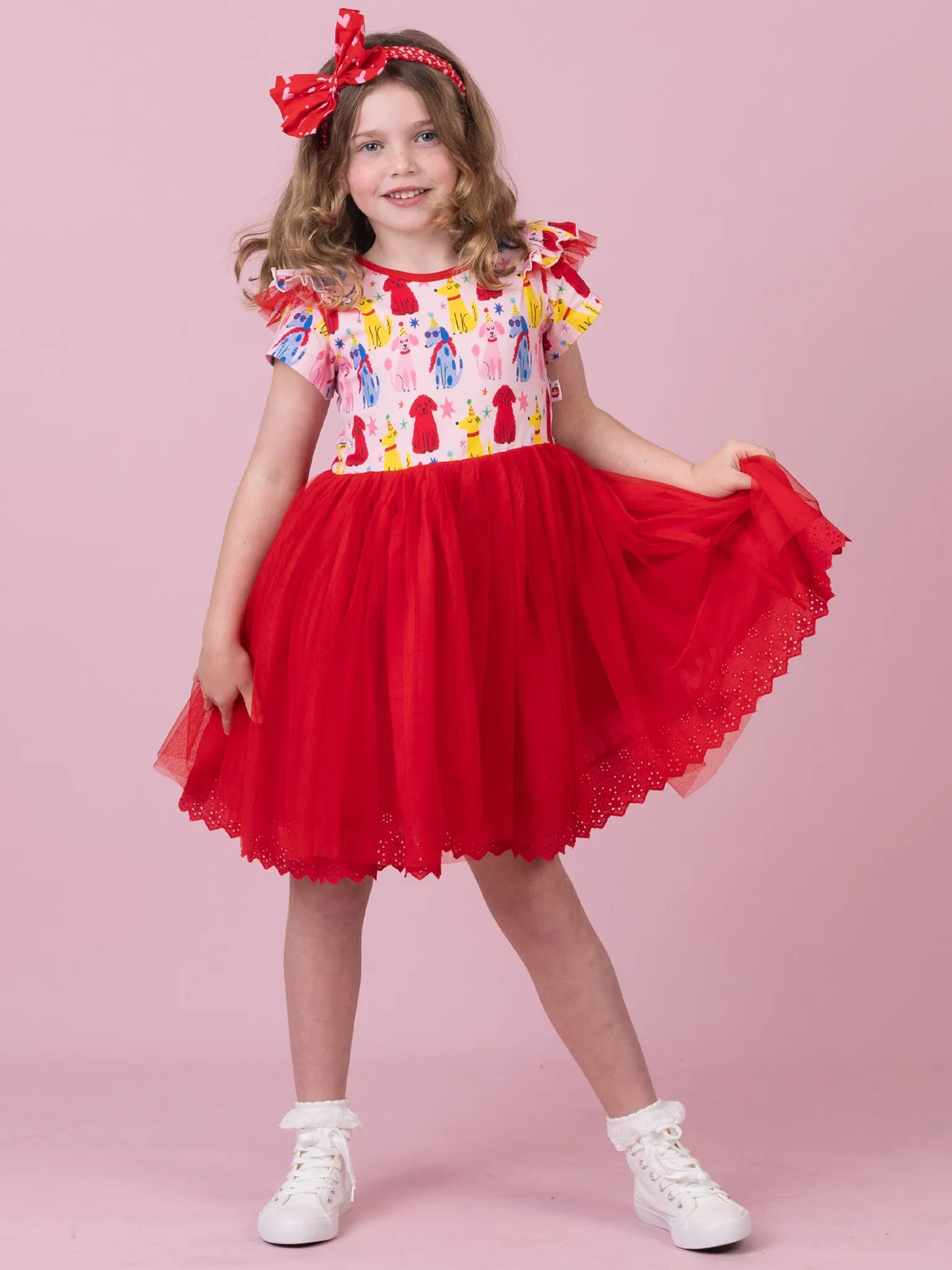 A young girl modeling a red and white dress with a colorful patterned bodice, red tulle skirt, and a red bow headband, standing against a solid pink studio background.