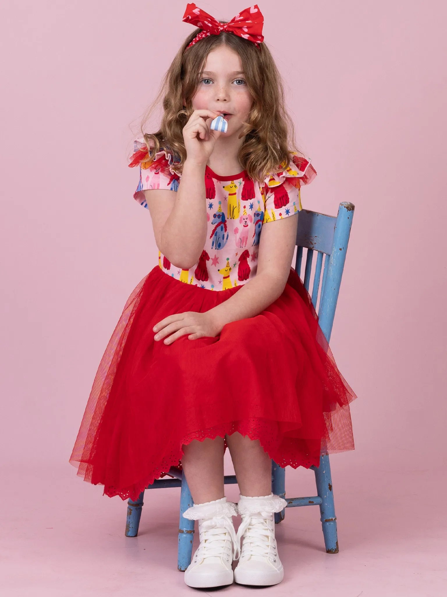 A young girl sits on a blue wooden chair against a solid pink studio background, wearing a red tulle skirt, a colorful patterned top, and a red bow headband.