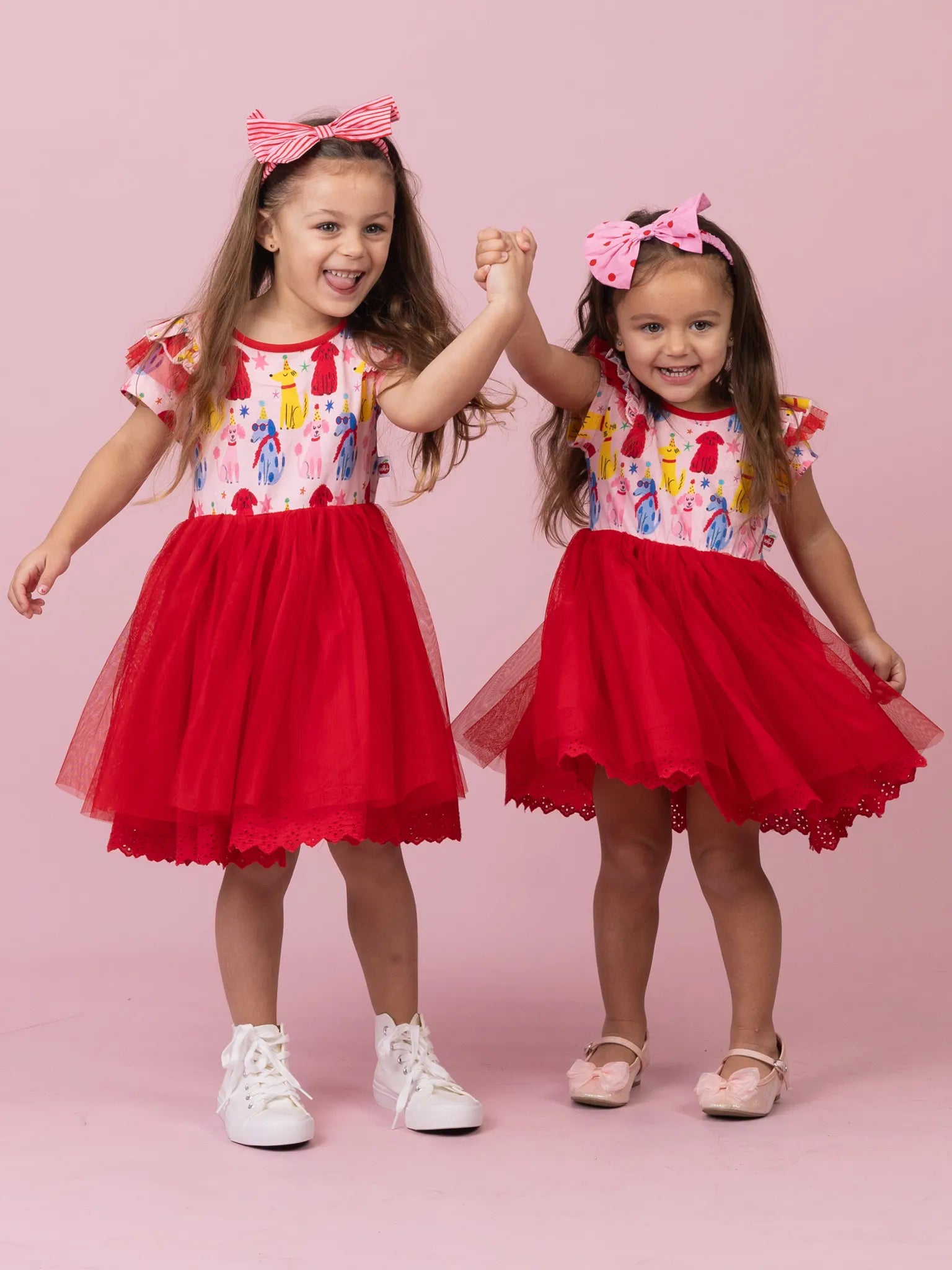 Two young girls wearing matching red tulle dresses with colorful ice cream-themed bodices and pink bow headbands, standing against a solid pink studio background.