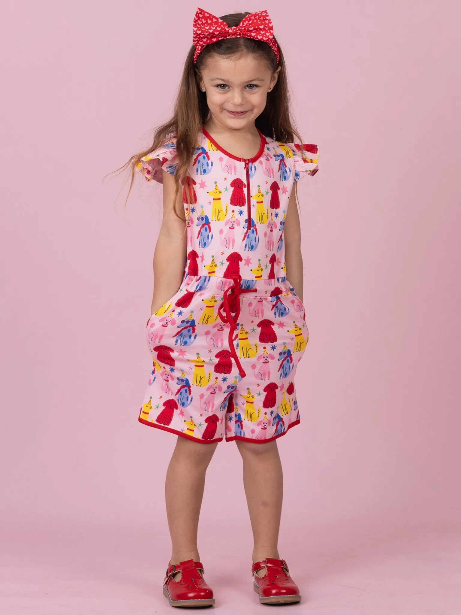 A young girl stands against a solid pink studio background wearing a short-sleeve romper with a colorful dog print, a red bow headband, and red shoes.